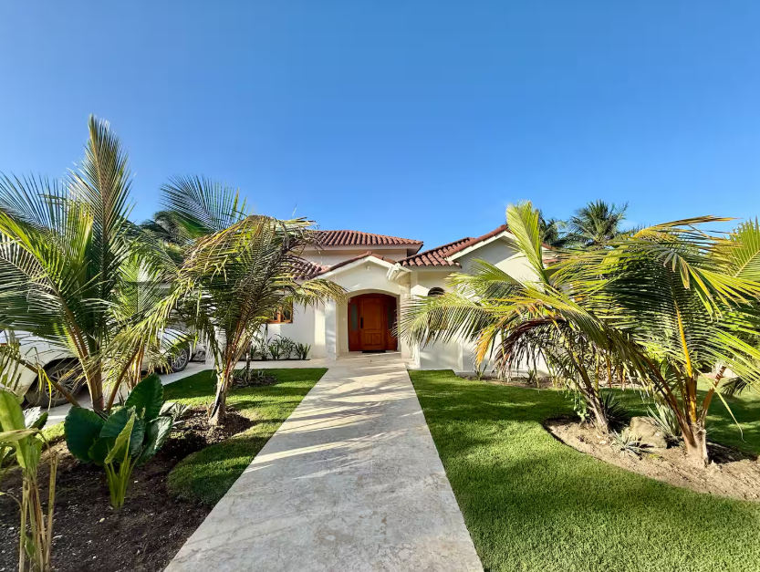 Front view of a house with a concrete pathway, surrounded by landscaped grass and palm trees, under a clear blue sky.