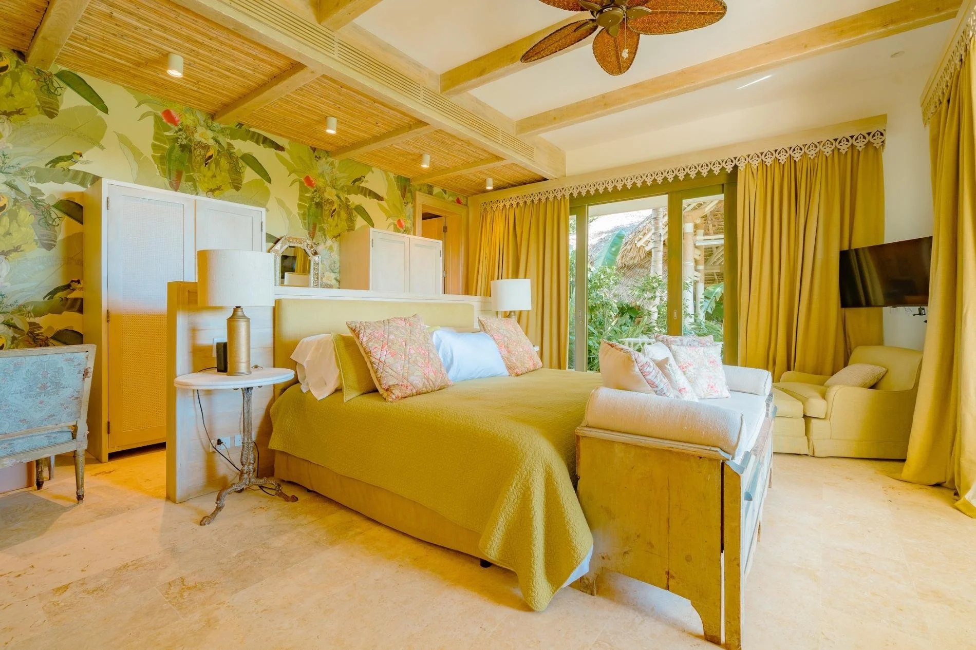 A cozy bedroom with tropical decor, yellow bedding, and multiple pillows. There are large windows with yellow curtains, a ceiling fan with leaf-shaped blades, and tropical plants visible outside. The room has wooden ceiling beams and natural light.
