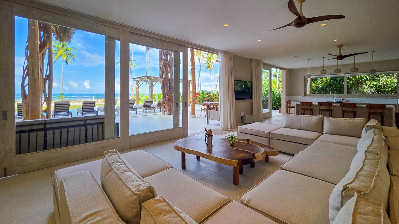 Living room with large glass sliding doors leading to a beachfront outdoor patio, featuring lounge chairs, palm trees, and the ocean in the background, with a white sectional sofa, wooden coffee table, and tropical decor.
