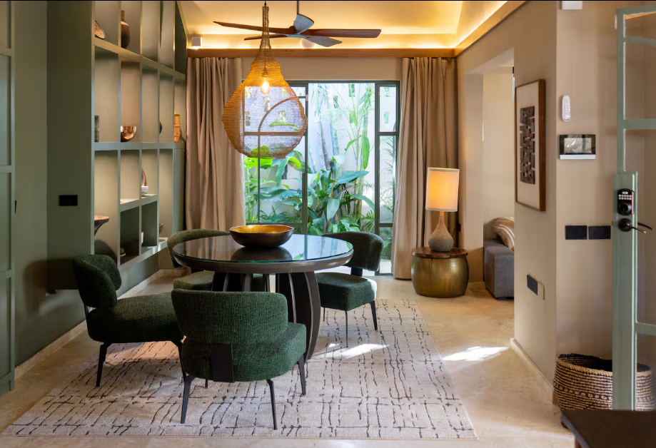 Living room with green accents, a round dining table with four green chairs, a large window with tropical plants outside, a decorative hanging light, a floor lamp, and artwork on the wall.