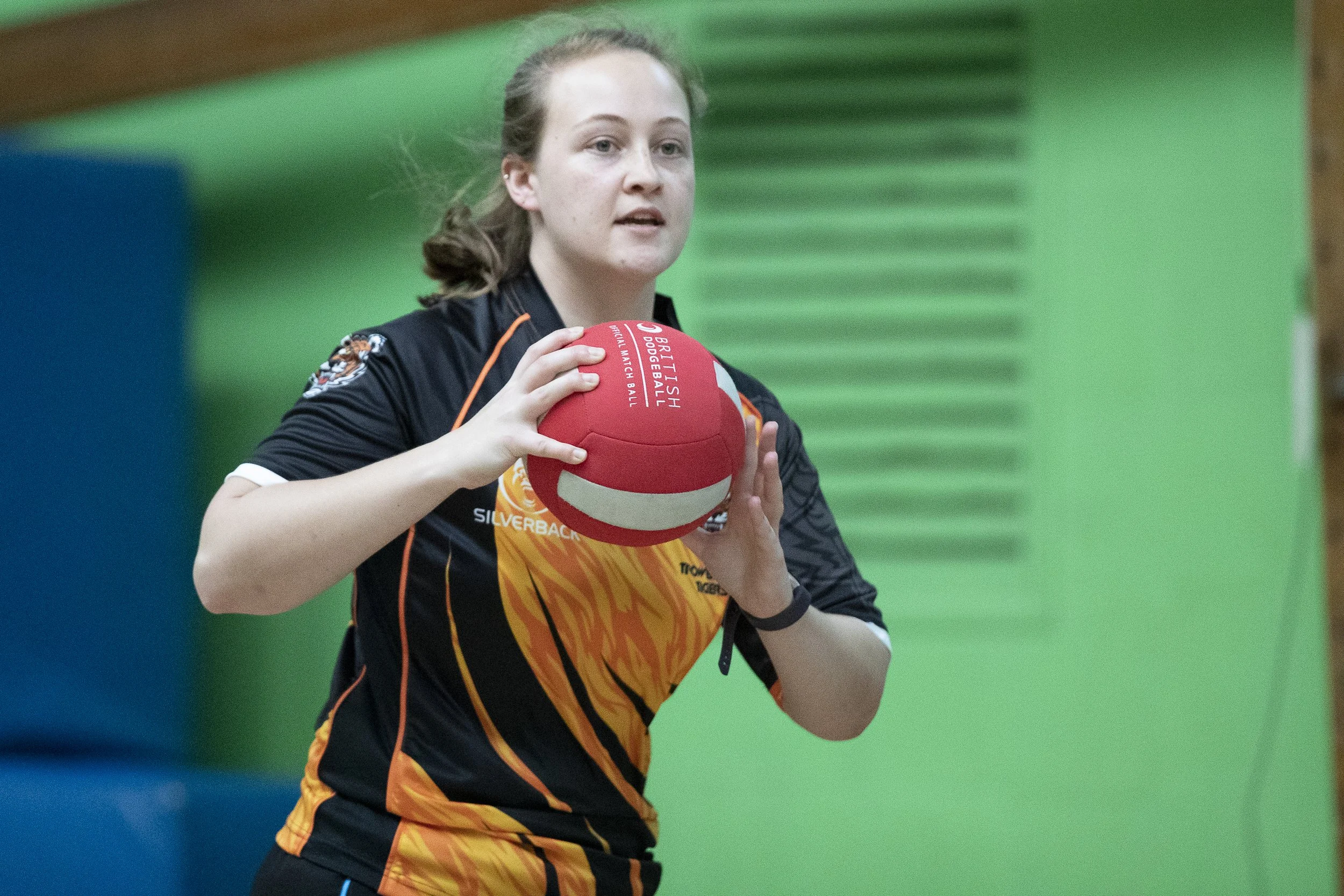 A young woman with light skin and brown hair tied back, wearing an Australian rules football uniform, holding a red and white football, during a match or practice in an indoor sports facility with green and blue padded walls.