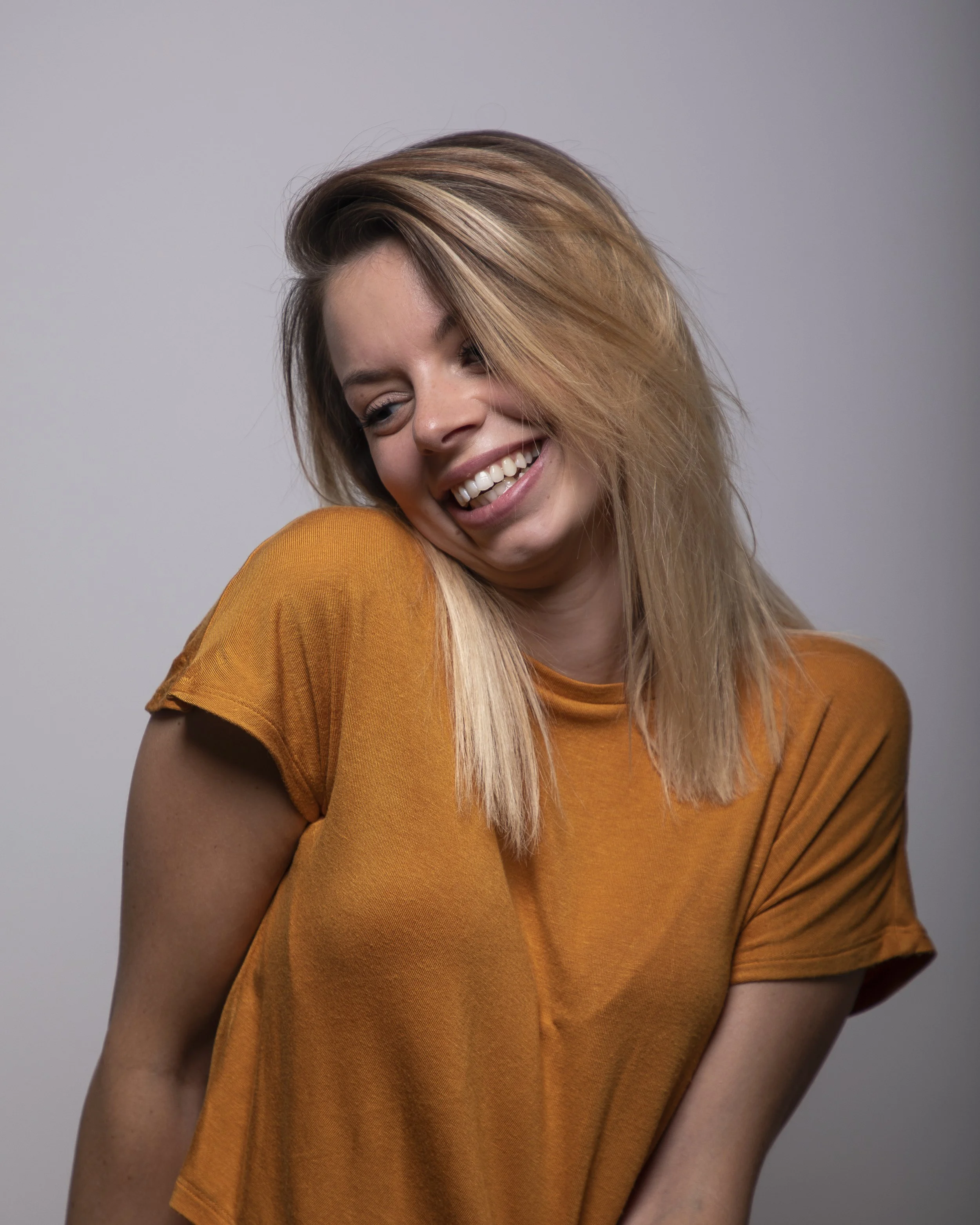 A young woman with long blonde hair styled to one side, smiling with eyes closed, wearing an orange T-shirt, against a plain gray background.