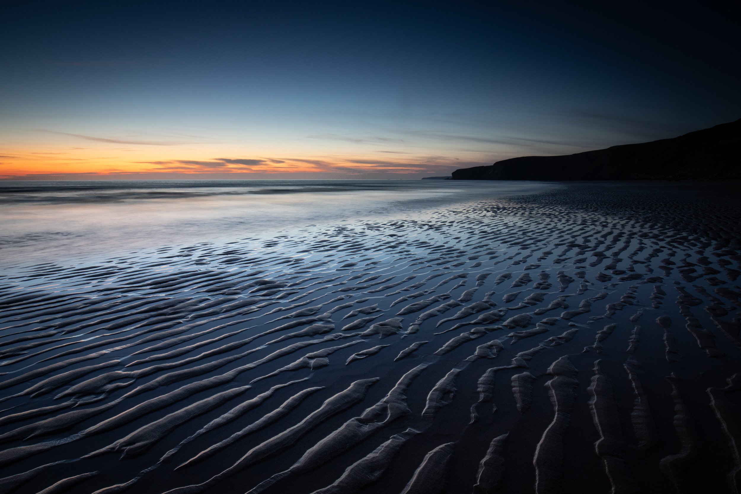 View of a beach during sunset with rippled sand and calm ocean waves, silhouette of cliffs in the distance, sky transitioning from orange to dark blue.