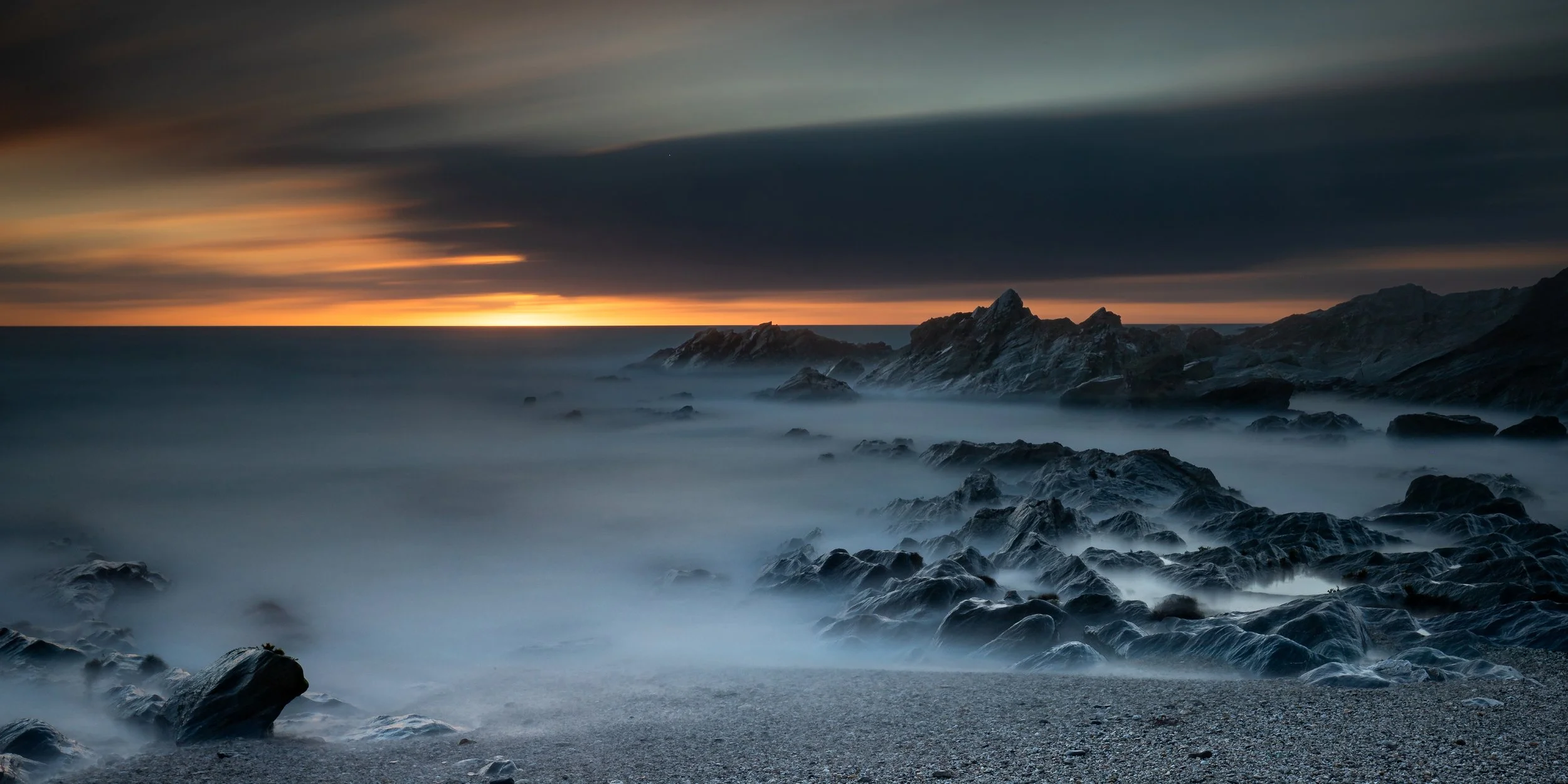 A rugged North Cornwall coastline at sunset, with rocky cliffs and foggy water, under a dark sky with orange and blue hues.