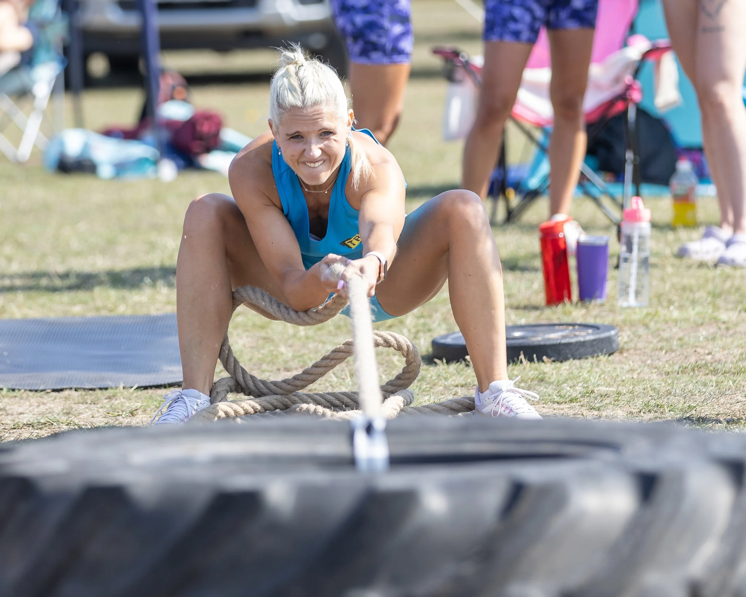 A woman with blonde hair tied up, wearing a blue athletic outfit, participating in an outdoor fitness event involving tug-of-war on a grassy field with various people and supplies in the background.