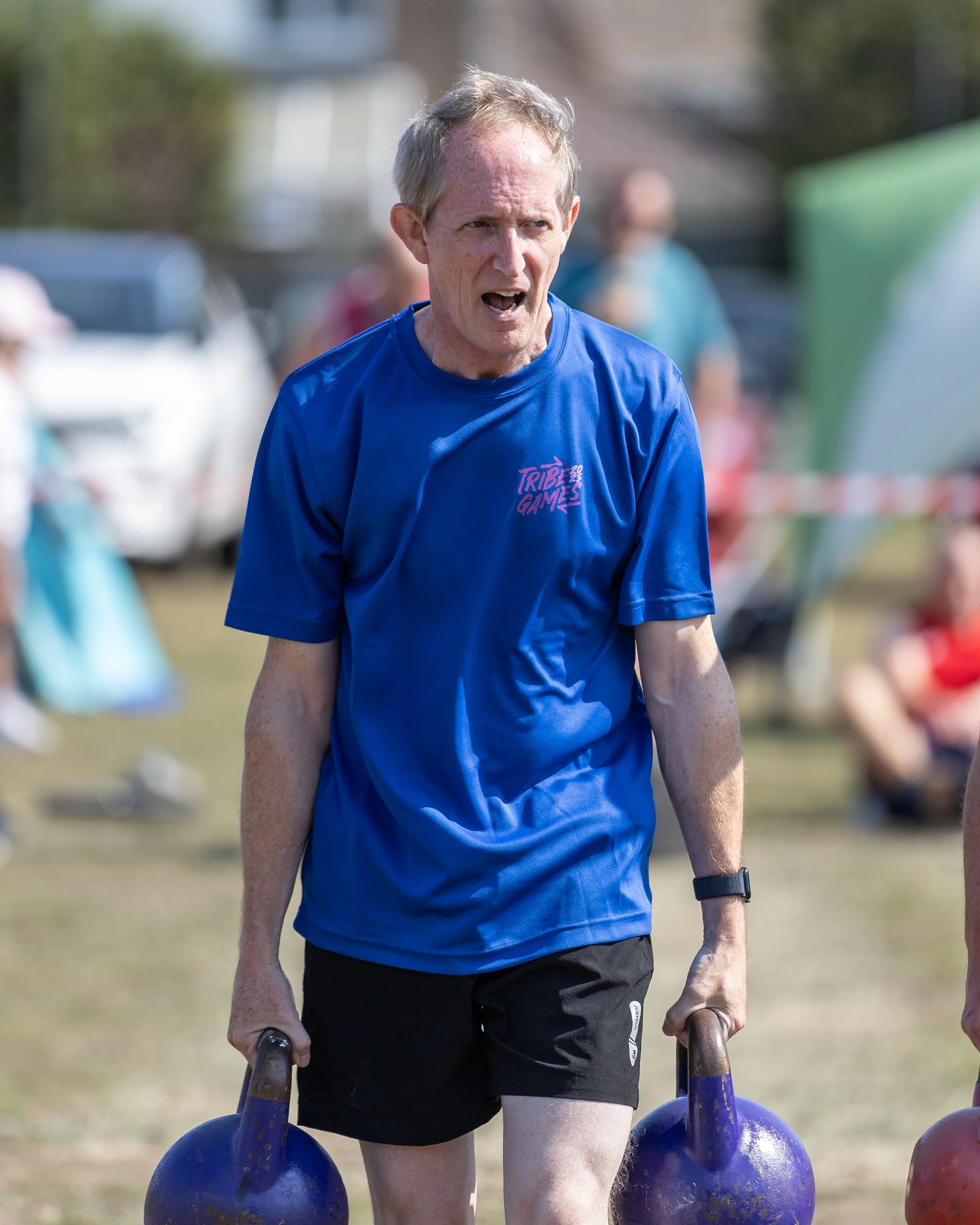 An older man in a blue t-shirt and black shorts is participating in an outdoor activity involving lifting heavy kettlebells. He appears to be focused and exerting effort. The background is slightly blurred, showing other people and outdoor elements.