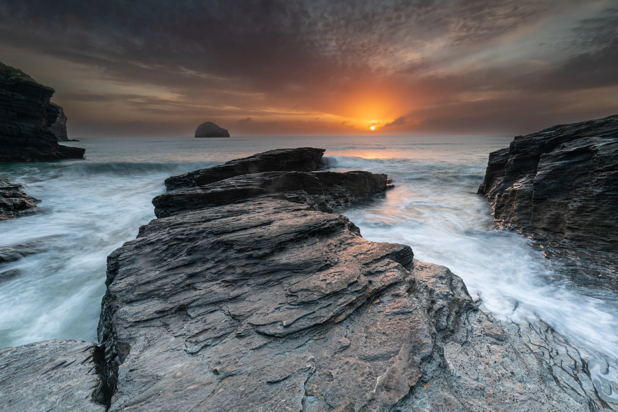 Sea rocks at sunset with clouds in the sky and the sun near the horizon over the ocean.
