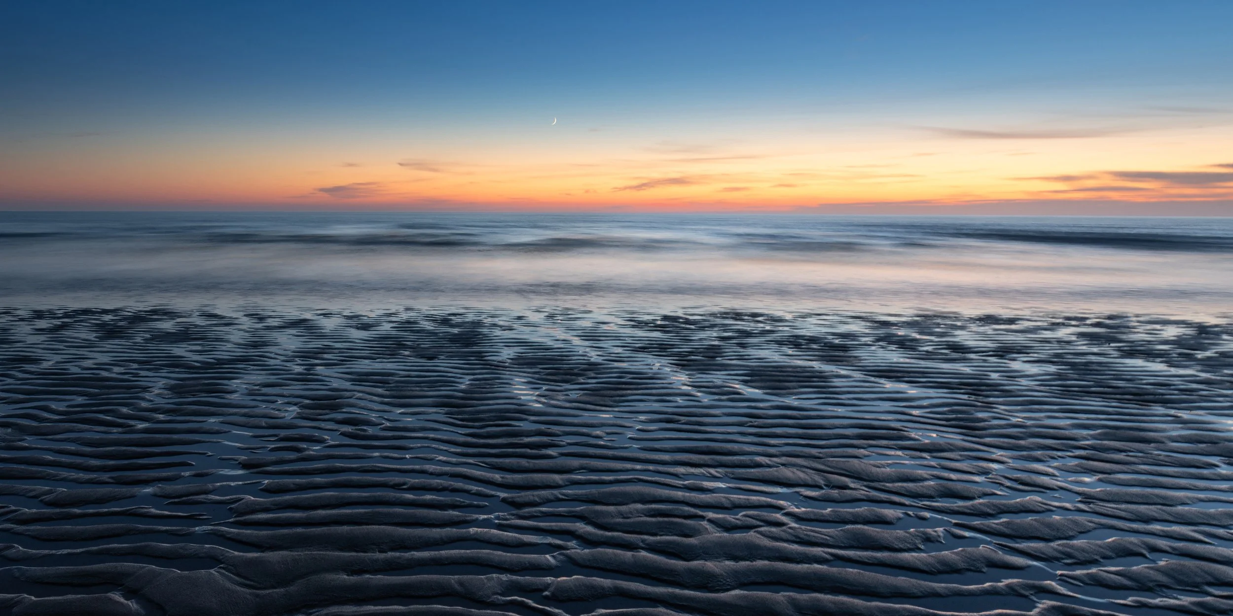 Beach scene at Watergate Bay, Cornwall, during sunset with sand patterns in the foreground, gentle waves in the middle, and a colorful sky with a crescent moon in the background.
