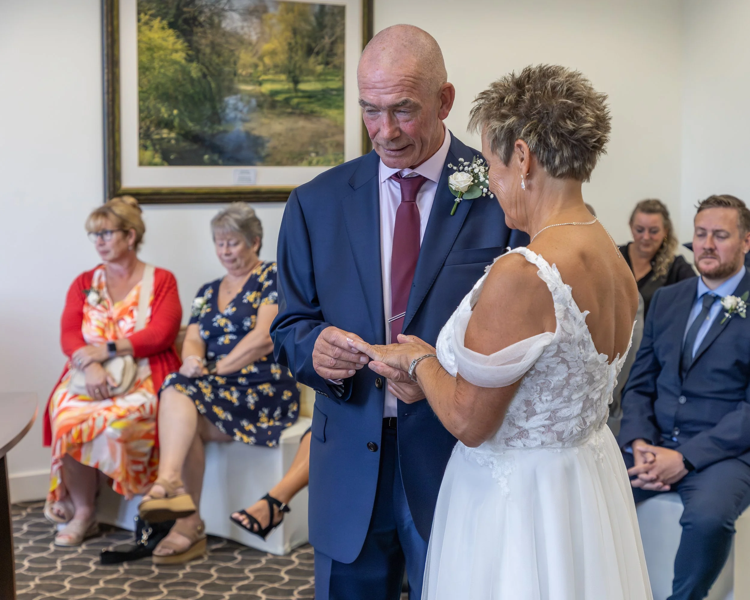 A groom in a navy suit with a white rose boutonniere is holding hands and looking down to an older woman in a white dress during a wedding ceremony, with guests seated in the background.
