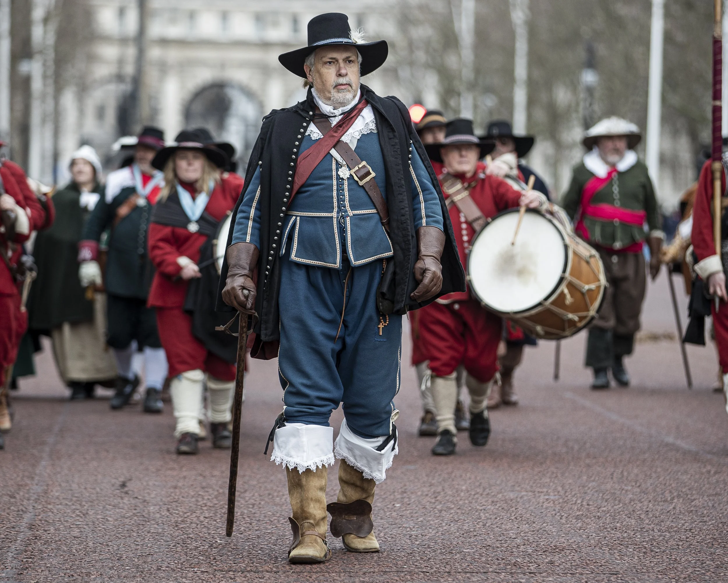 A man dressed in 18th-century colonial attire leads a parade in a historical reenactment. The man wears a large black hat with a feather, a blue coat with white trim, lace cuffs, and leather gloves, and carries a walking stick. Behind him, women and 
