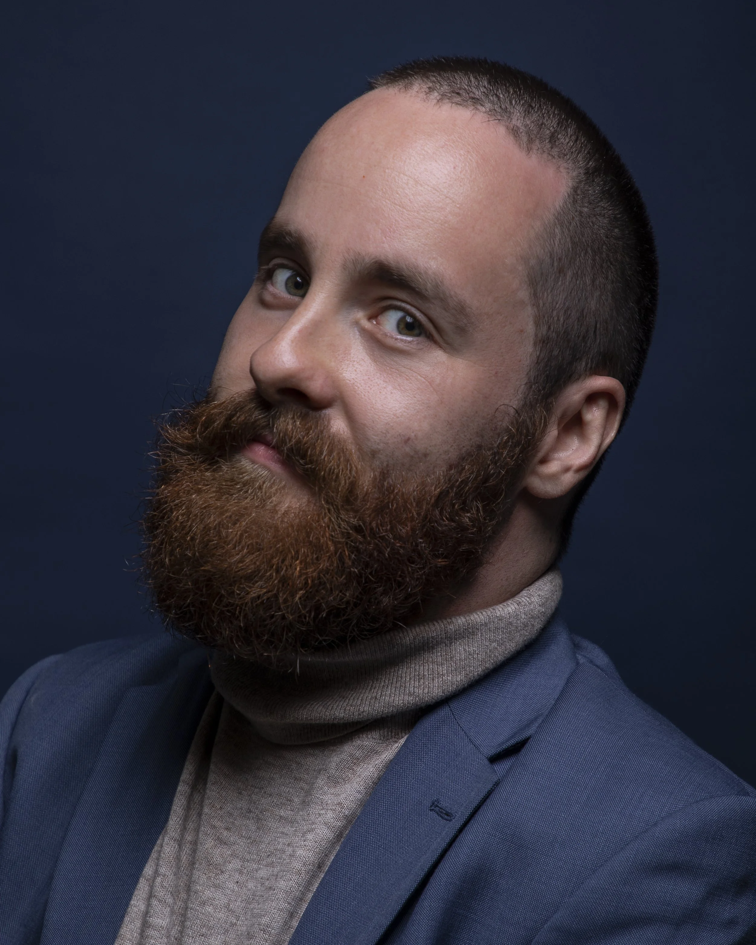 Close-up of a man with a beard, wearing a blue blazer and a gray turtleneck, against a dark background.