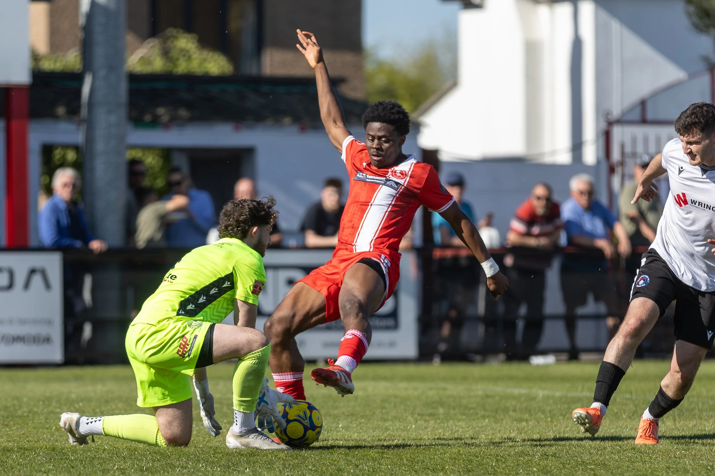 A soccer match with a player in a red uniform jumping over the goalkeeper in a neon yellow uniform, who is on one knee on the ground, trying to block a soccer ball. Other players and spectators are visible in the background.