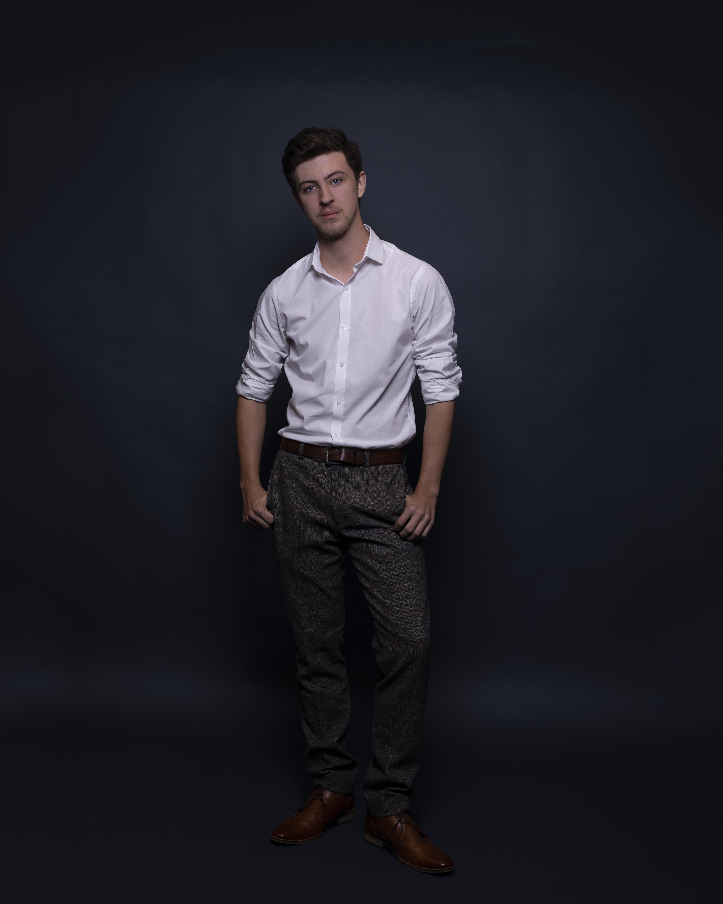 Young man in white dress shirt and grey pants standing against dark background with relaxed pose