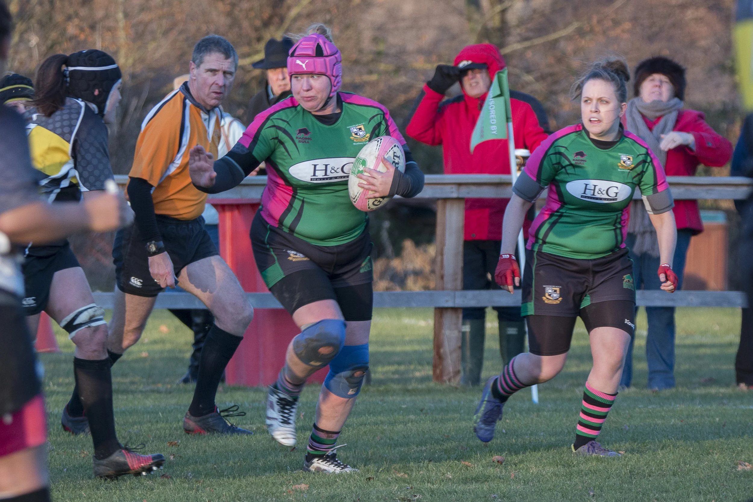 Women playing rugby on a grassy field with spectators watching in the background.