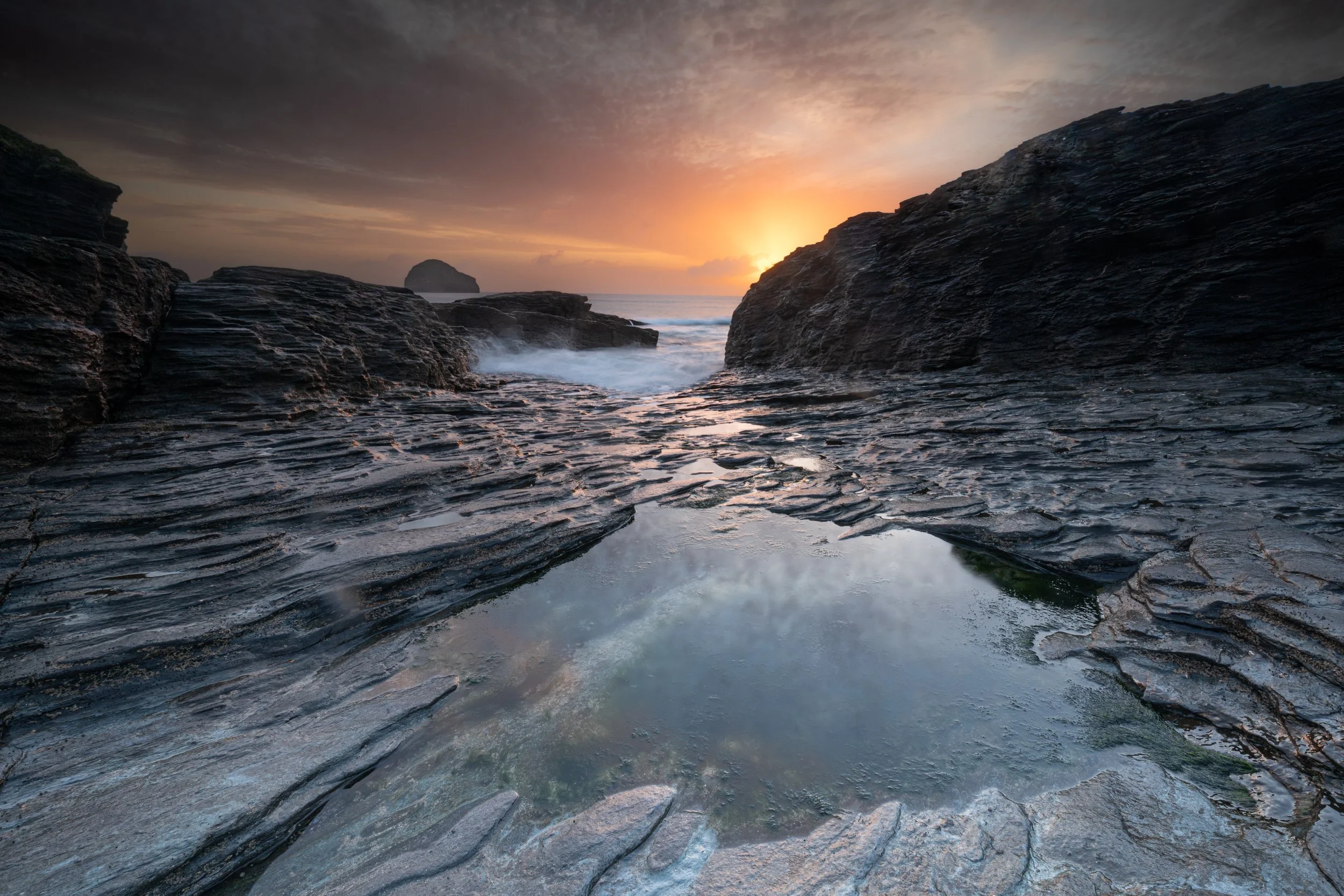 A rocky coastline at sunset with dark, textured rocks in the foreground, a larger rock formation in the background, and a sunset sky with clouds and warm orange and yellow hues.