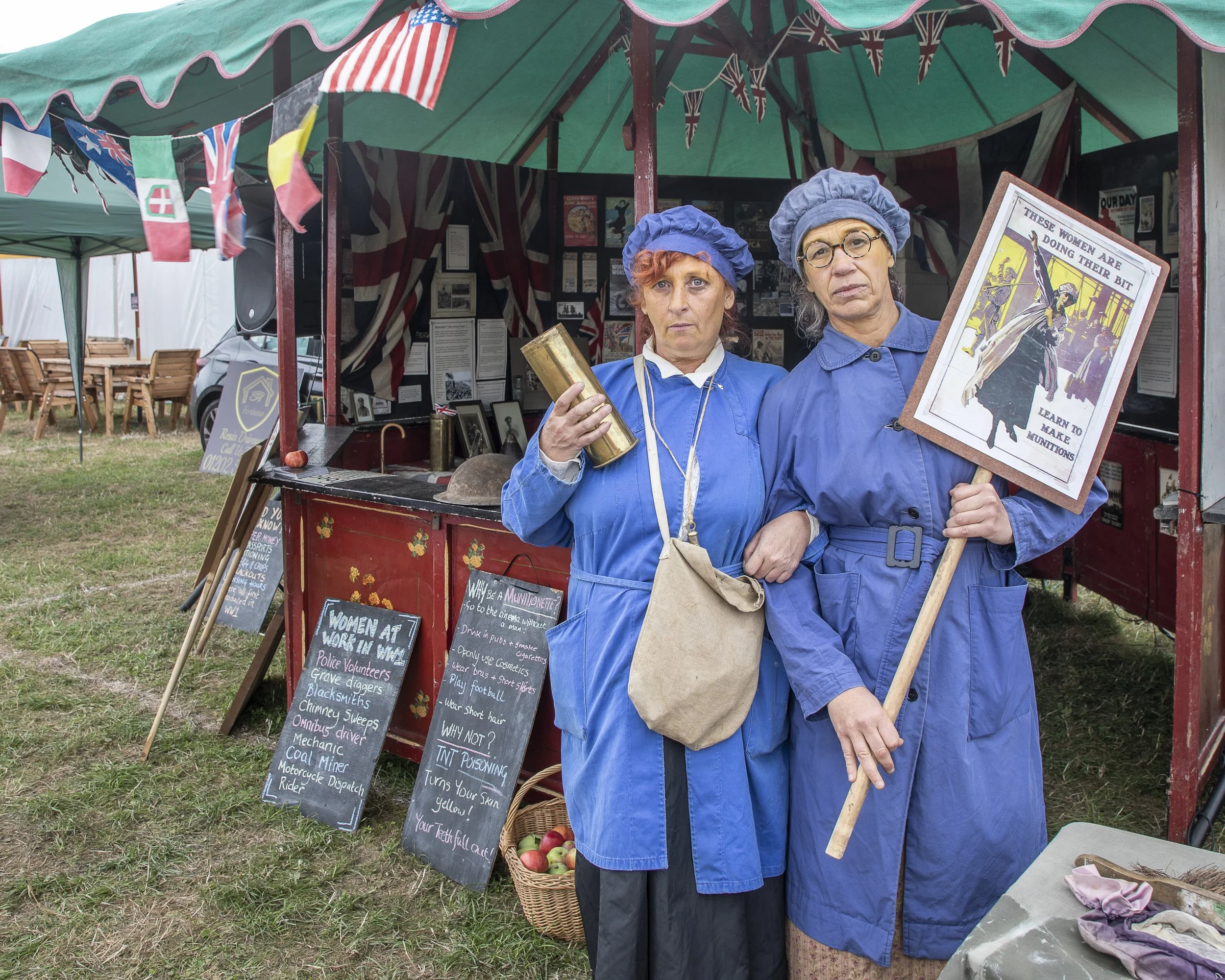 Two women dressed as nurses in blue uniforms and nurse caps stand inside a vintage-style booth decorated with Union Jack flags and bunting at an outdoor event. One woman holds a framed poster about women doing their bit during wartime, and the other 
