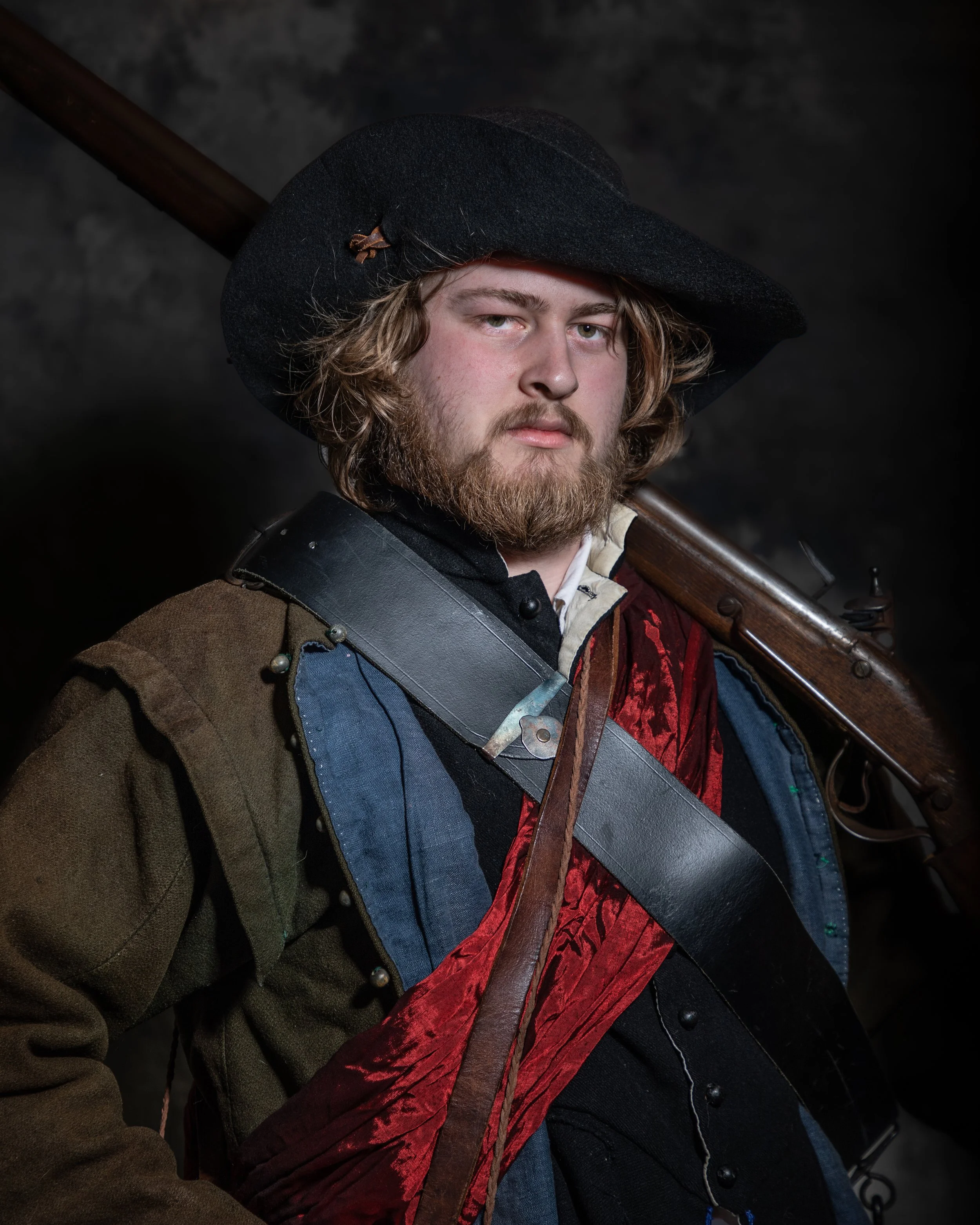 A man with a beard and shoulder-length curly hair wearing a large black hat, a brown coat, and a red and blue medieval-style garment. He is holding a long rifle or musket over his shoulder and looking directly at the camera against a dark background.