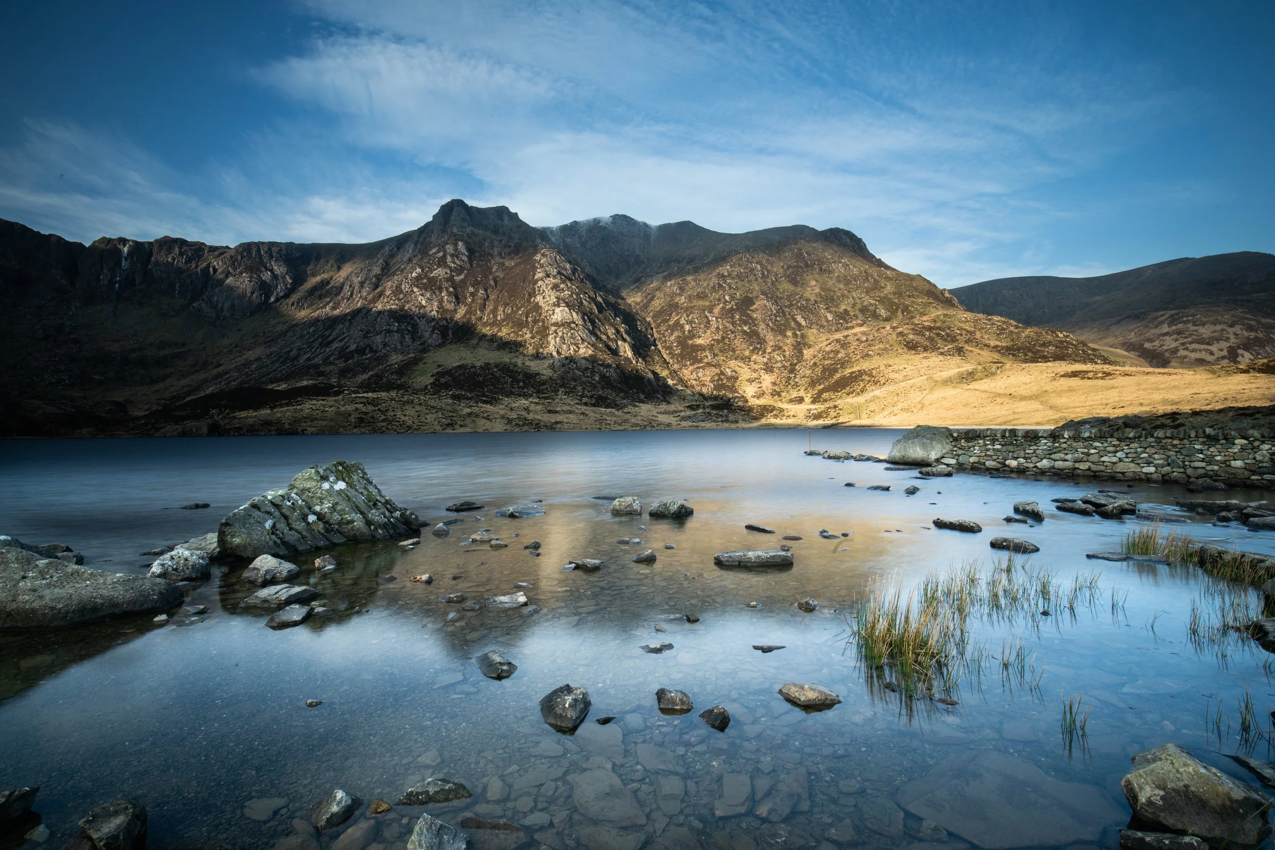 A scenic landscape featuring a calm lake at the base of rugged mountains with a partly cloudy sky overhead. Llyn Idwal, Snowdonia, North Wales.