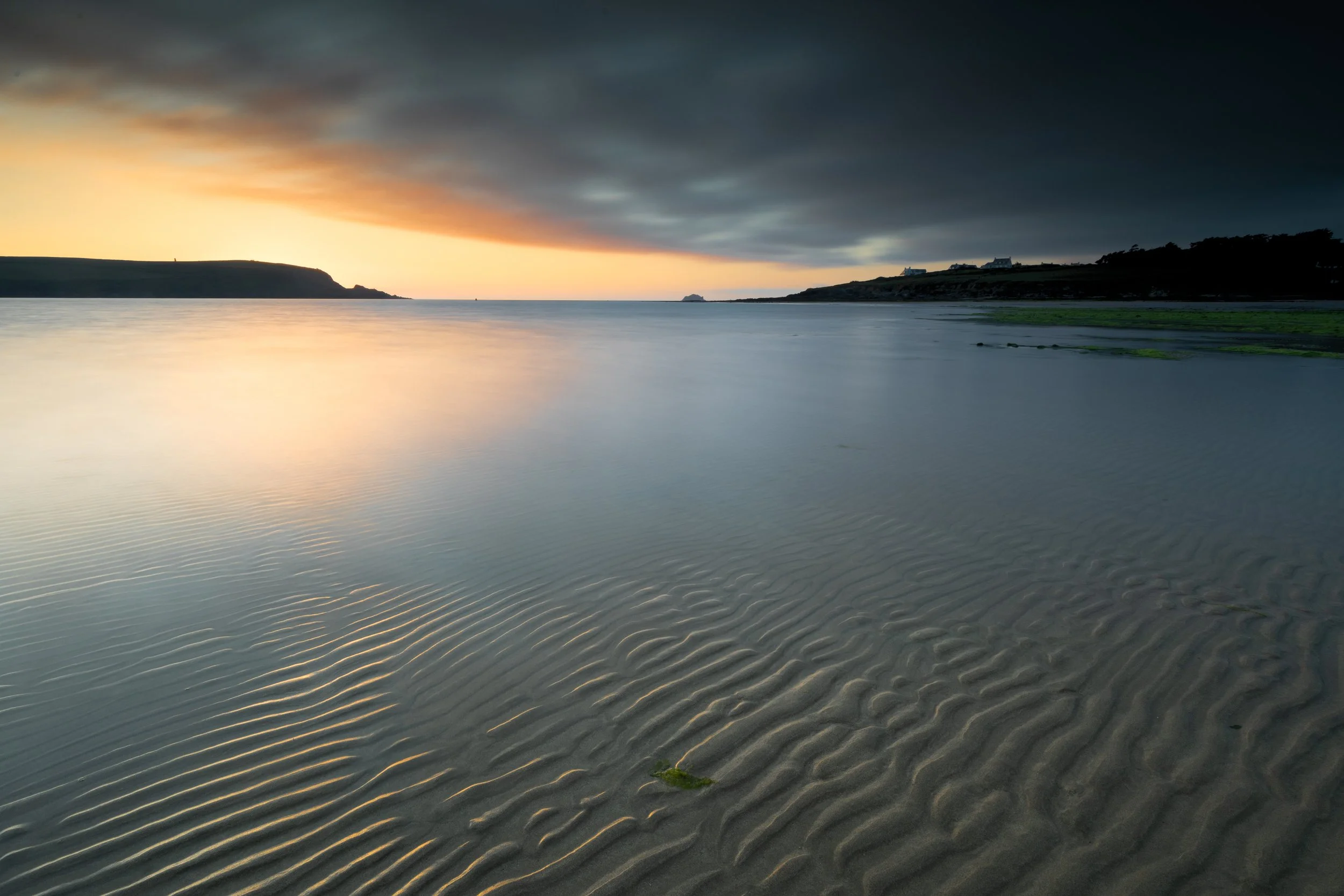 A serene coastal scene at sunset with calm water, rocky shoreline, and overcast sky.