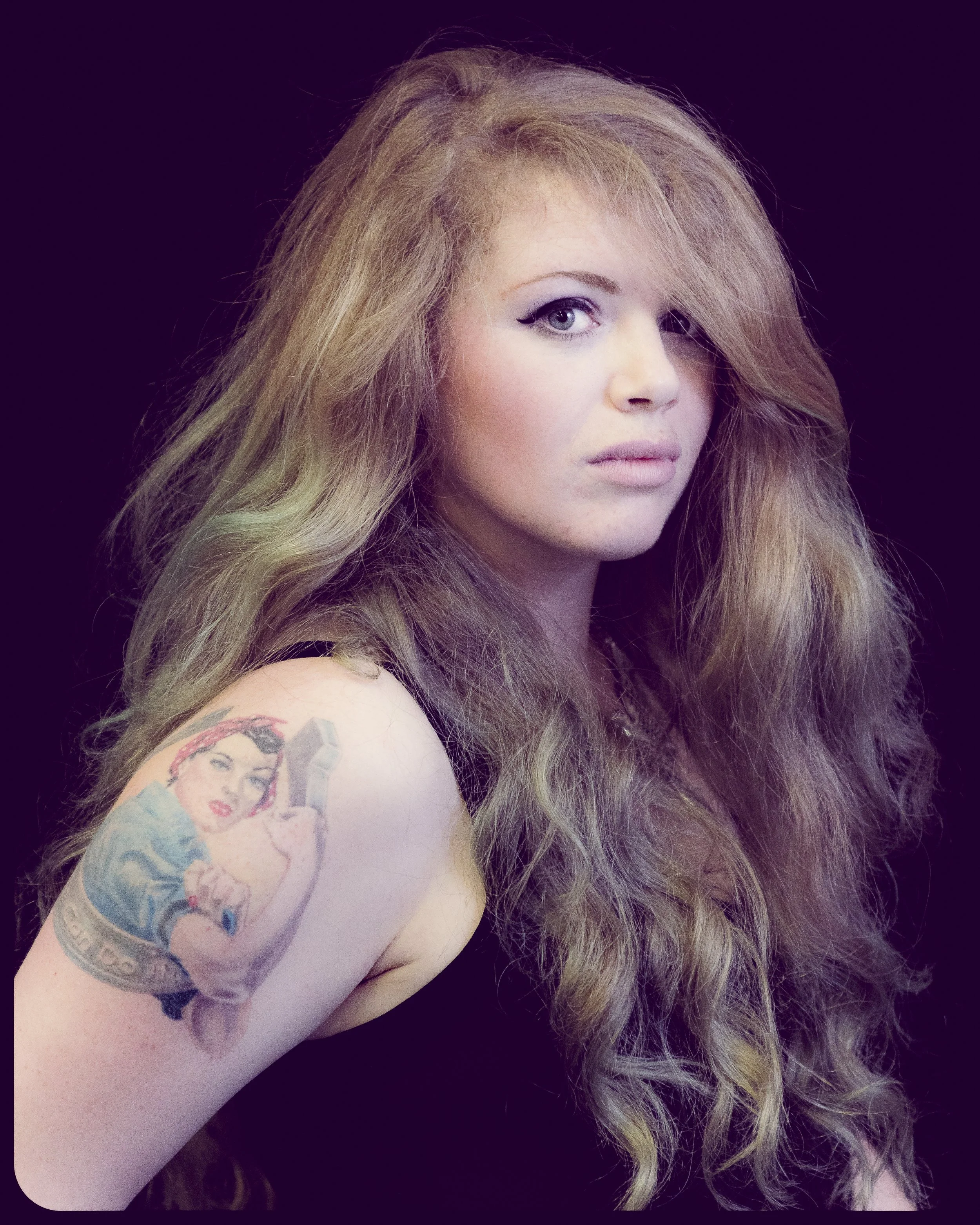 A woman with long, wavy blonde hair and eye makeup, wearing a sleeveless top, showing a colorful tattoo of a woman's face on her upper arm, against a dark background.