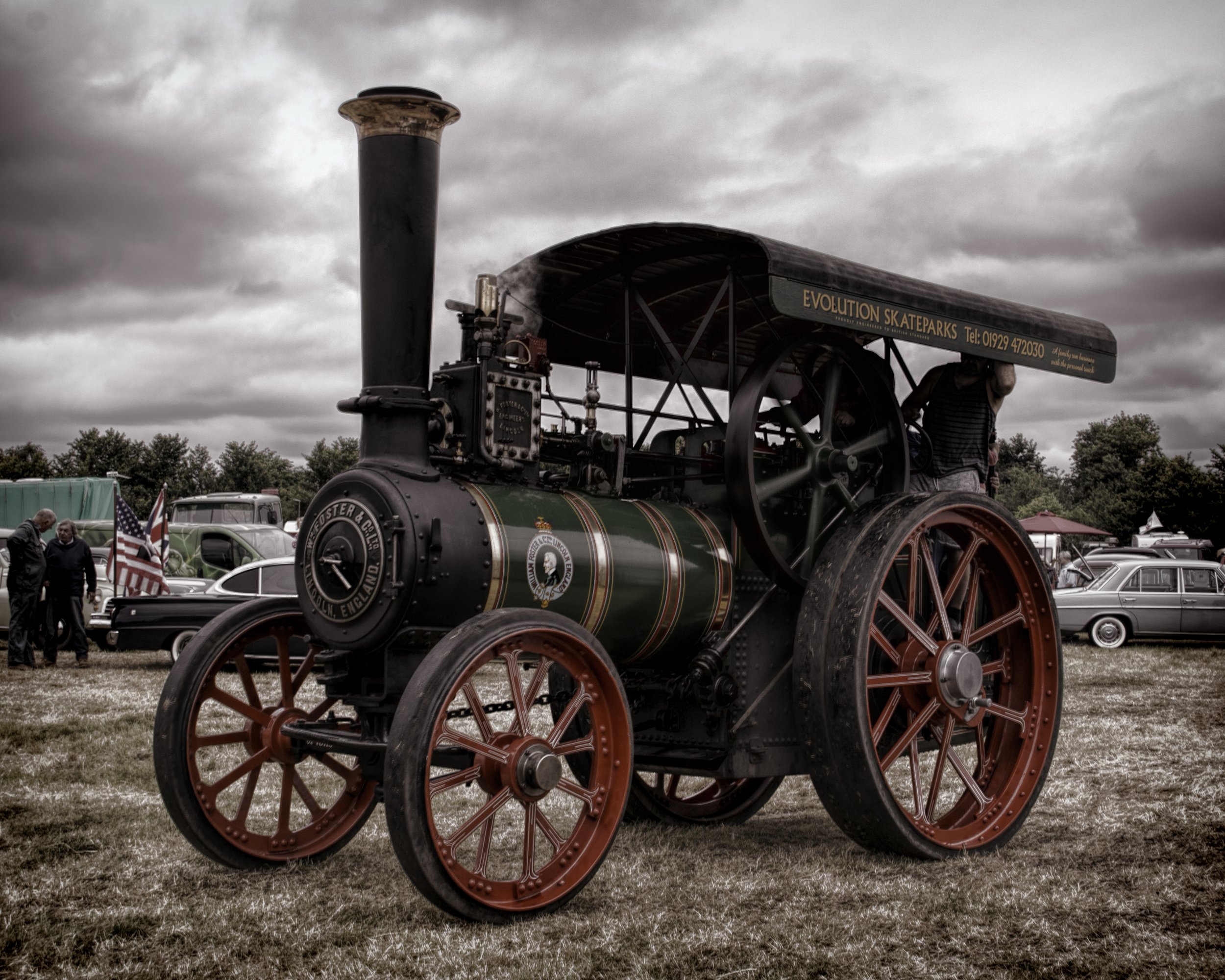 A vintage steam-powered traction engine with large red wheels and a tall black chimney, displayed outdoors on a grassy field during a cloudy day, with people and cars visible in the background.