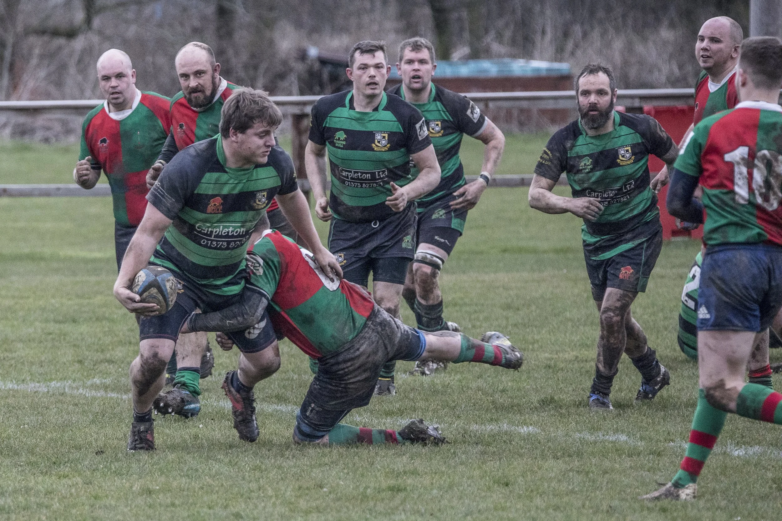 A rugby match with players in green and black stripes and in red and green. One player is on the ground holding the ball, while others run or prepare to tackle on a muddy field.
