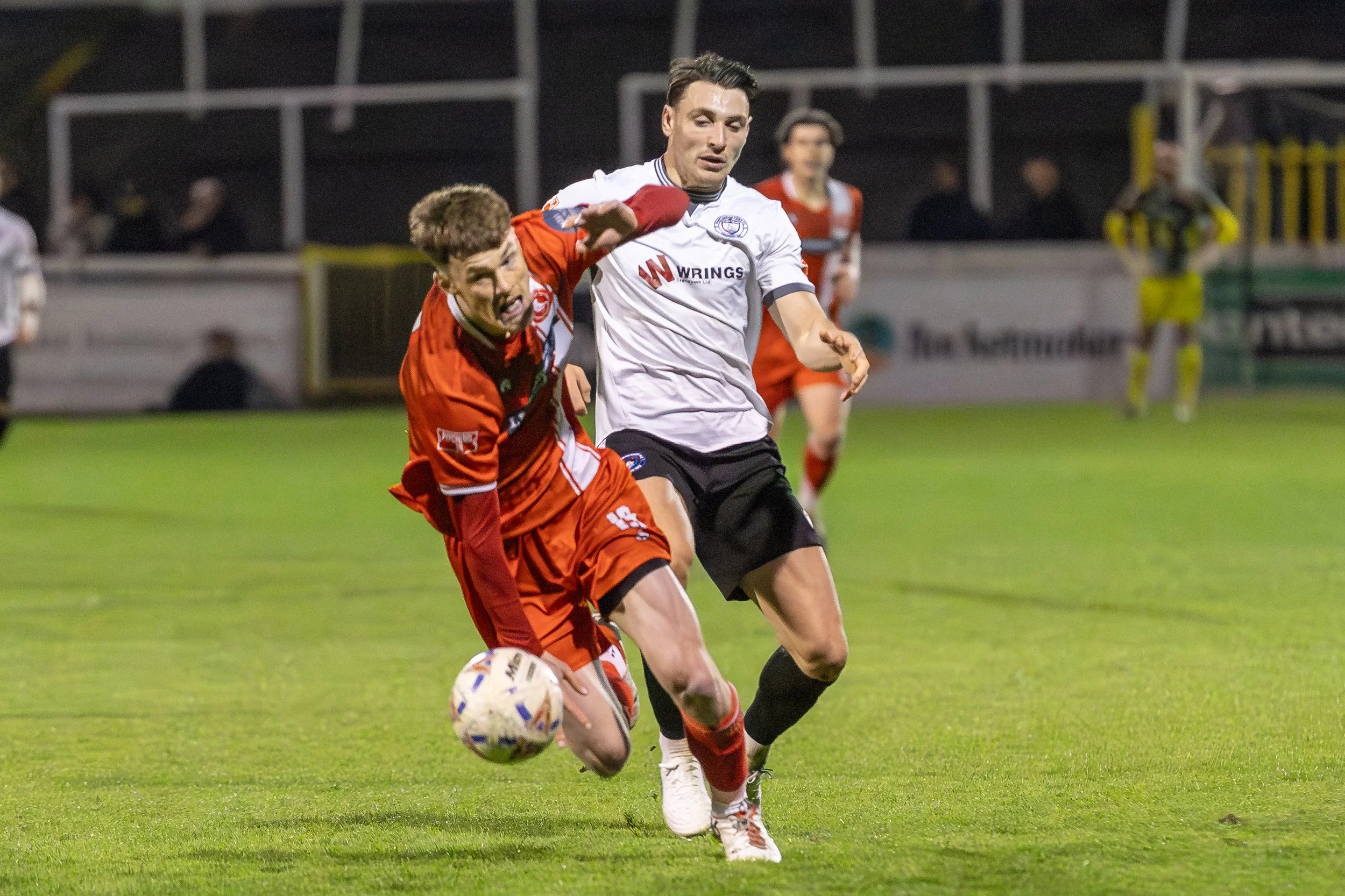 Two soccer players are competing for possession of the ball on a field during a night game. One player is wearing a red uniform and the other is wearing a white jersey with black shorts.