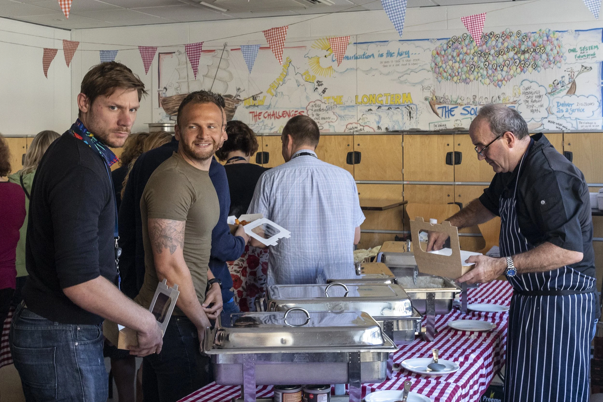 A group of people standing in line at a buffet table during an indoor event, with a colorful, hand-drawn mural on the wall behind them and red, white, and blue checkered and gingham bunting hanging overhead.