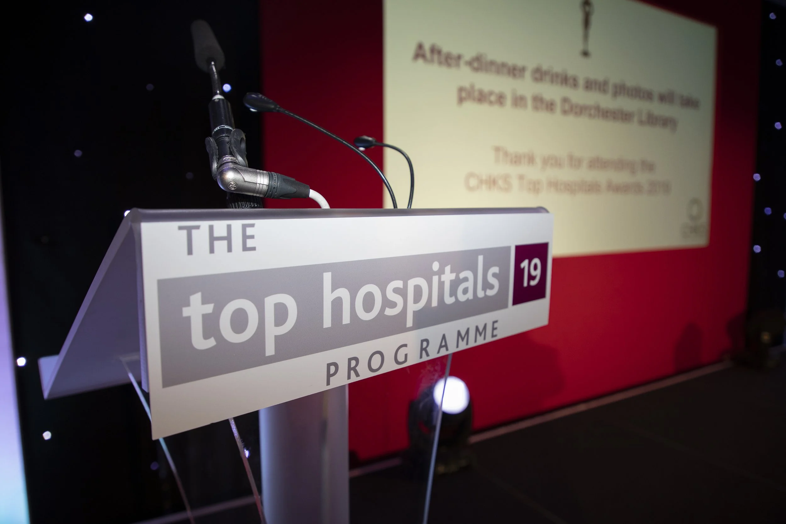 An event stage featuring a podium with a sign for a hospital and a microphone. In the background, there is a projected slide with partially visible text about an after-dinner drinks and an appreciation message for the CHKS Tag Hospital Awards 2019.