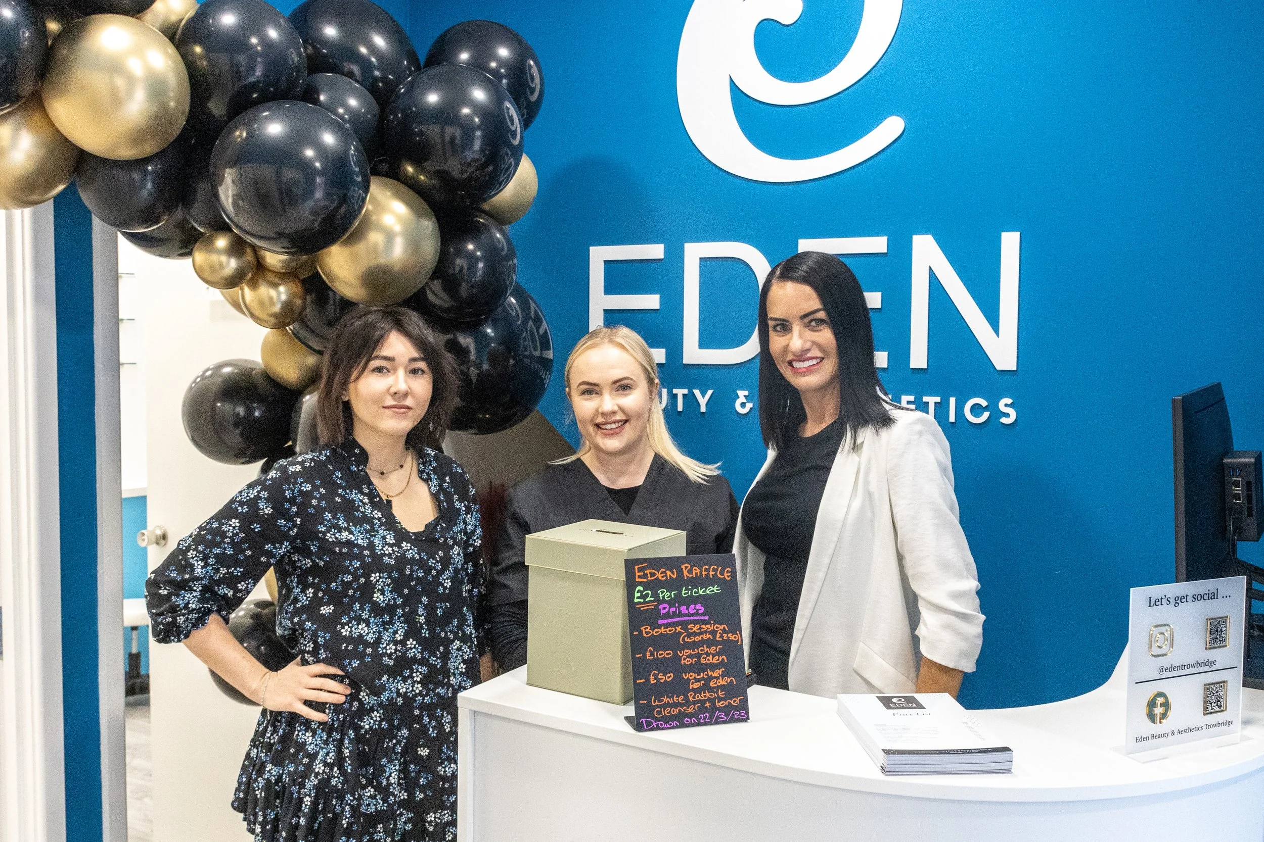 Three women stand behind a white counter in front of a blue wall with the words 'EDEN Beauty & Aesthetics' on it. One woman holds a box labeled 'Eden Raffle'. There are black and gold balloons on the side, and a colorful sign with raffle prizes on th