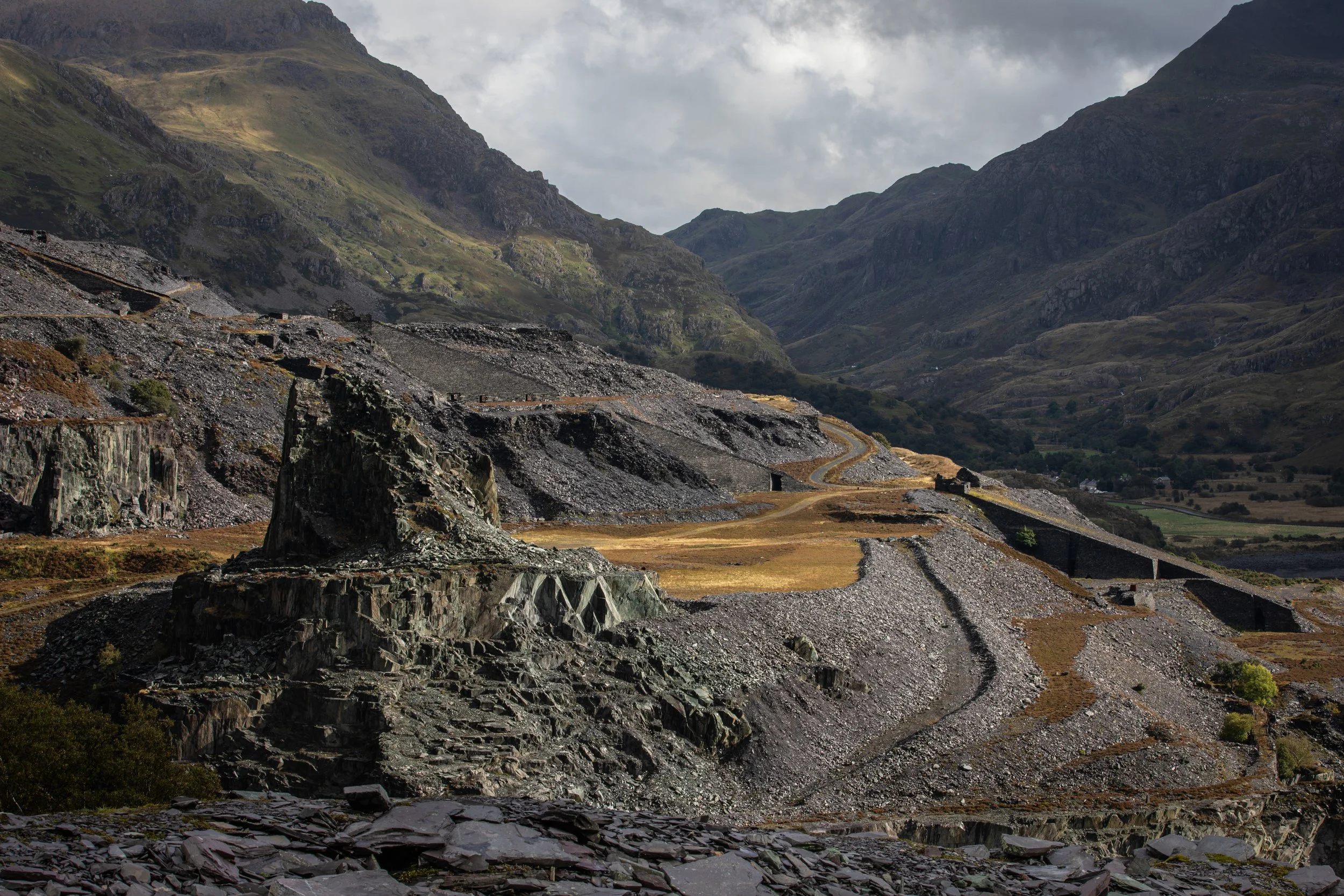 Scenic view of rugged mountains with rocky terrain, green patches, and a winding road amidst valley. Overcast sky with clouds and mist. Dinorwich Quarry, Snowdonia, North Wales.