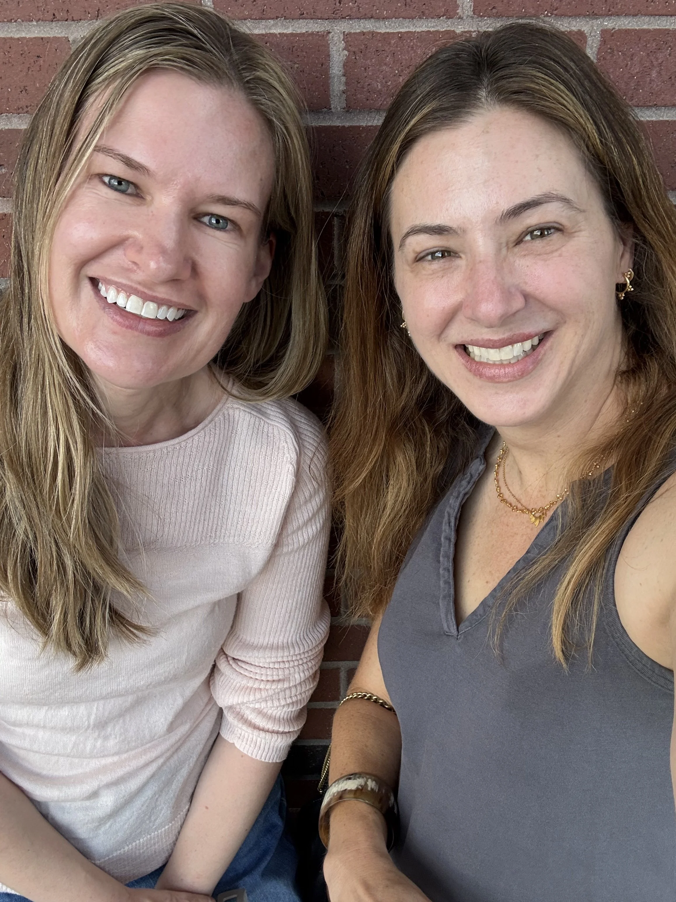 Close-up photo of two smiling women sitting next to each other against a brick wall, with visible jewelry and casual clothing.