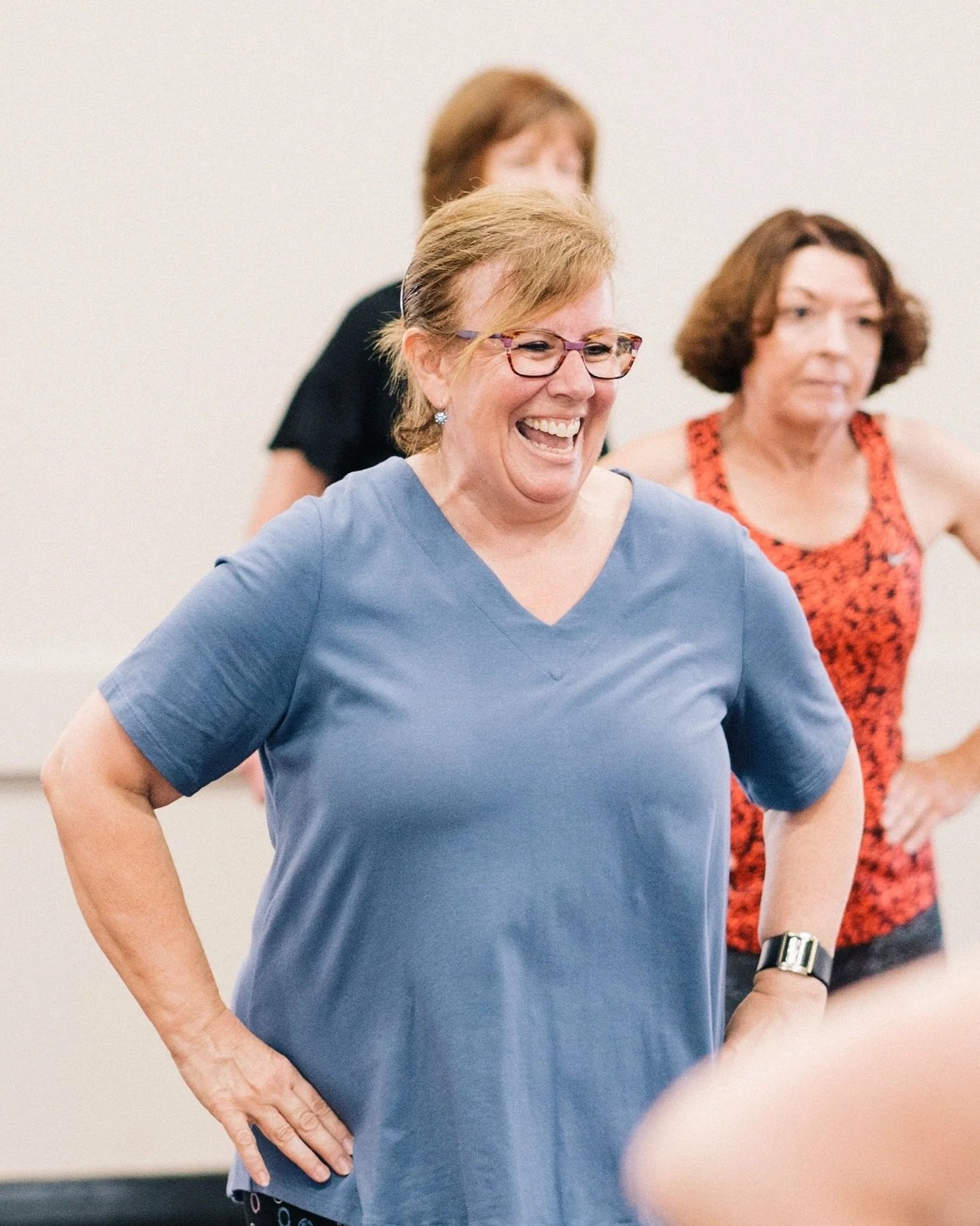 A middle-aged woman with short light brown hair, glasses, and earrings, is smiling and looking happy while standing with her hands on her hips in a casual setting. She is wearing a blue t-shirt and a watch on her left wrist. In the background, two other women are slightly out of focus, one wearing a black shirt and the other wearing a red patterned top.