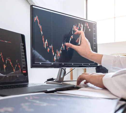 Person analyzing a stock market chart on a large monitor and a laptop in an office.