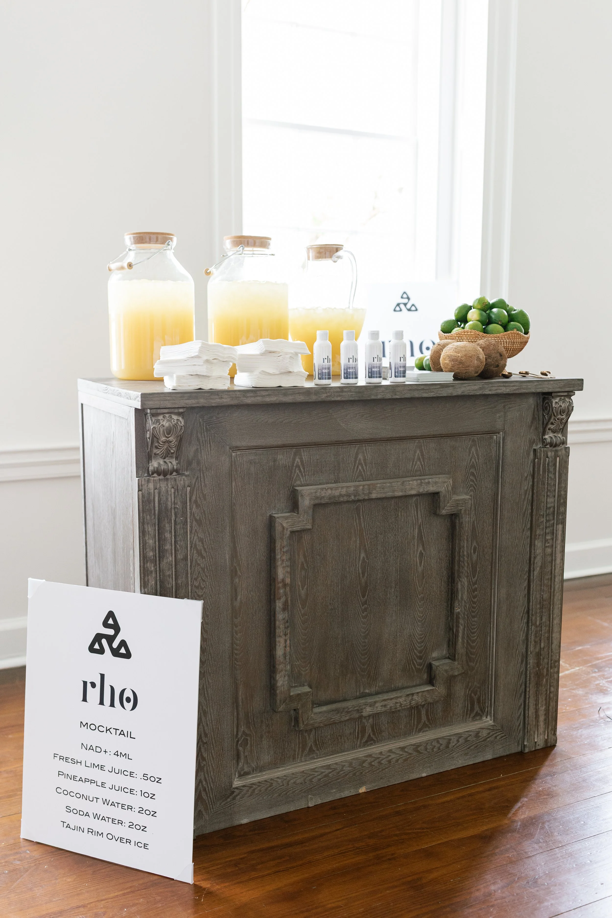 A wooden desk with three jars of pale yellow liquid, a pile of white napkins, four small bottles, and a bowl of green limes and coconuts. There is an A4-size white sign with black text showing a cocktail recipe in front of the desk and a white wall with a window behind it.