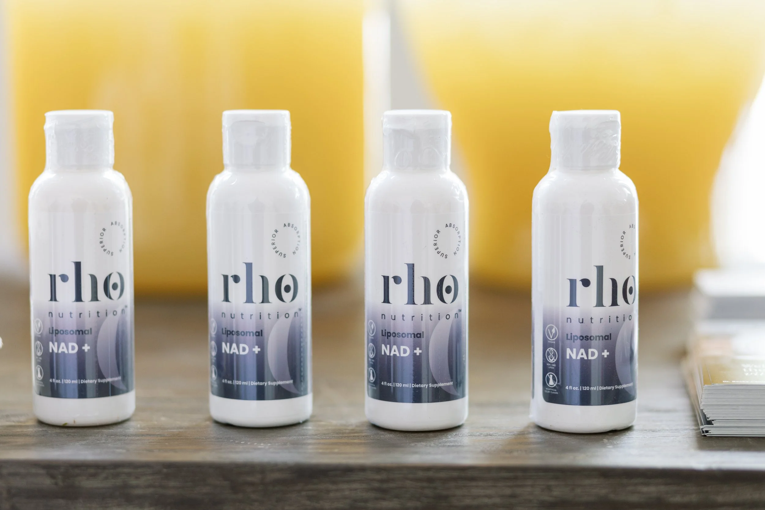 Four white bottles labeled 'the nutrition Liposomal NAD+' arranged in a row on a wooden surface with a yellow background.