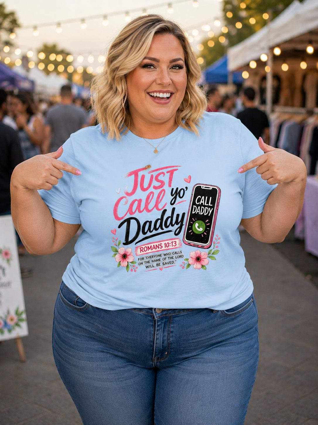 Woman standing outdoors at a market, wearing a light blue t-shirt with the words 'Just Call Yo Daddy' and an illustration of a smartphone with a call screen. She is pointing to her shirt, smiling, and has shoulder-length blonde hair. In the background, there are market stalls and string lights.