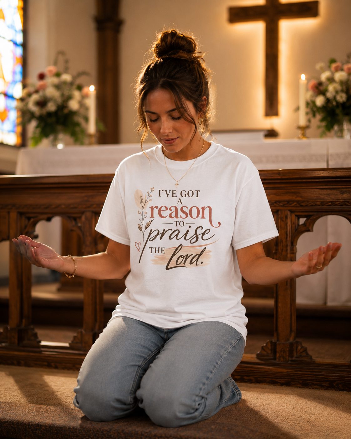 A woman kneeling in a church, praying with her eyes closed and hands open. She is wearing a white t-shirt that says 'I've got a reason to praise the Lord,' and has a cross necklace. The church has wooden pews, floral arrangements, candles, a large cross on the wall, and stained glass windows.