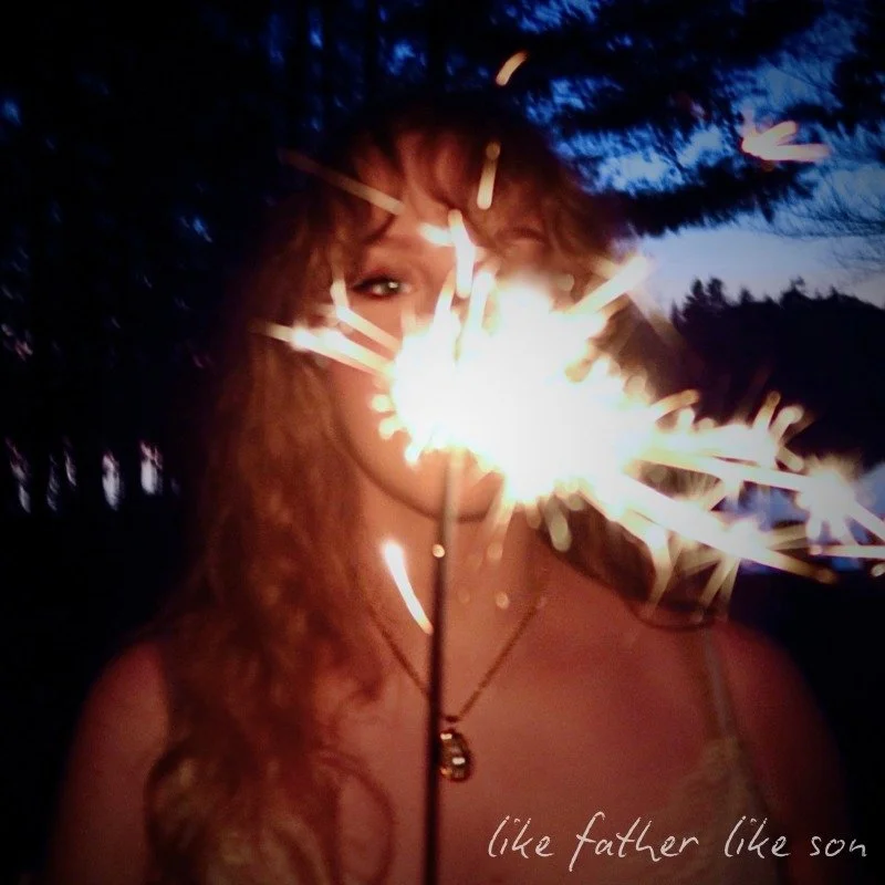 A woman outdoors at dusk holding a lit sparkler in front of her face, with trees in the background, and the words "like father like son" written at the bottom.