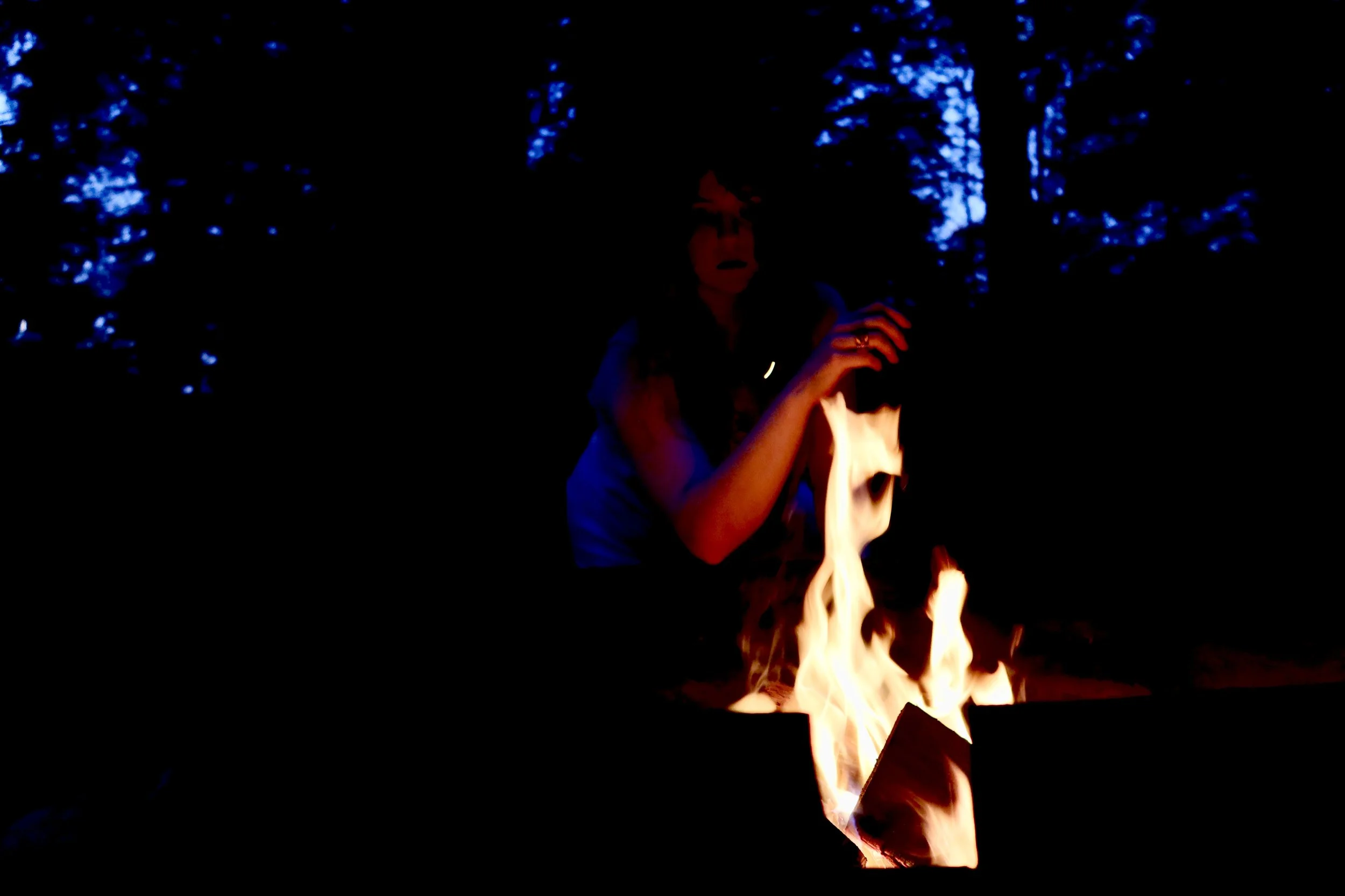 Person holding a paper in front of an open flame outdoors at night surrounded by dark trees.