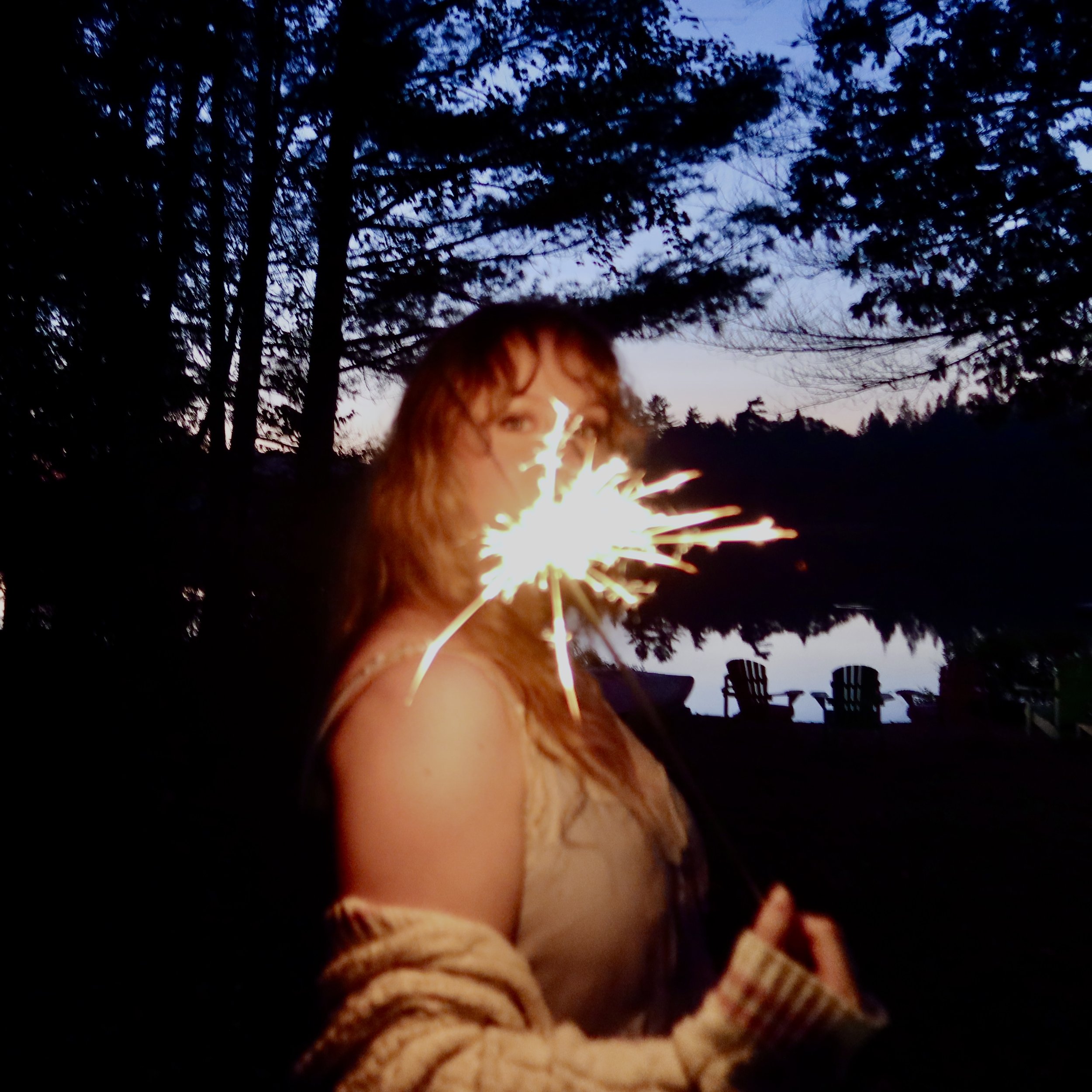 A person with reddish-brown hair and a beige sweater holding sparklers outdoors during dusk, with trees and blue sky in the background.