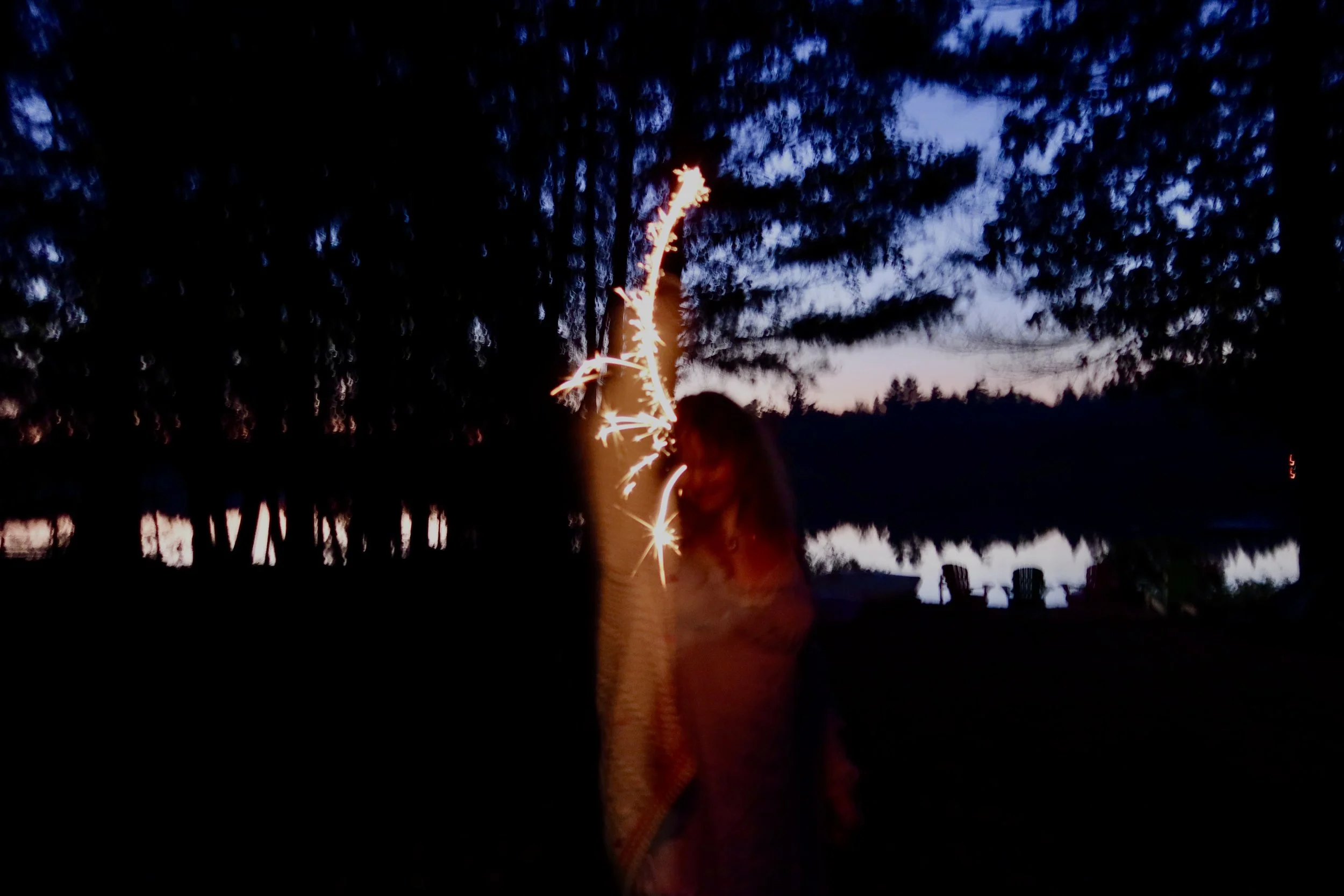 A person holding a lit sparkler during dusk near a body of water, with trees and chairs visible in the background.