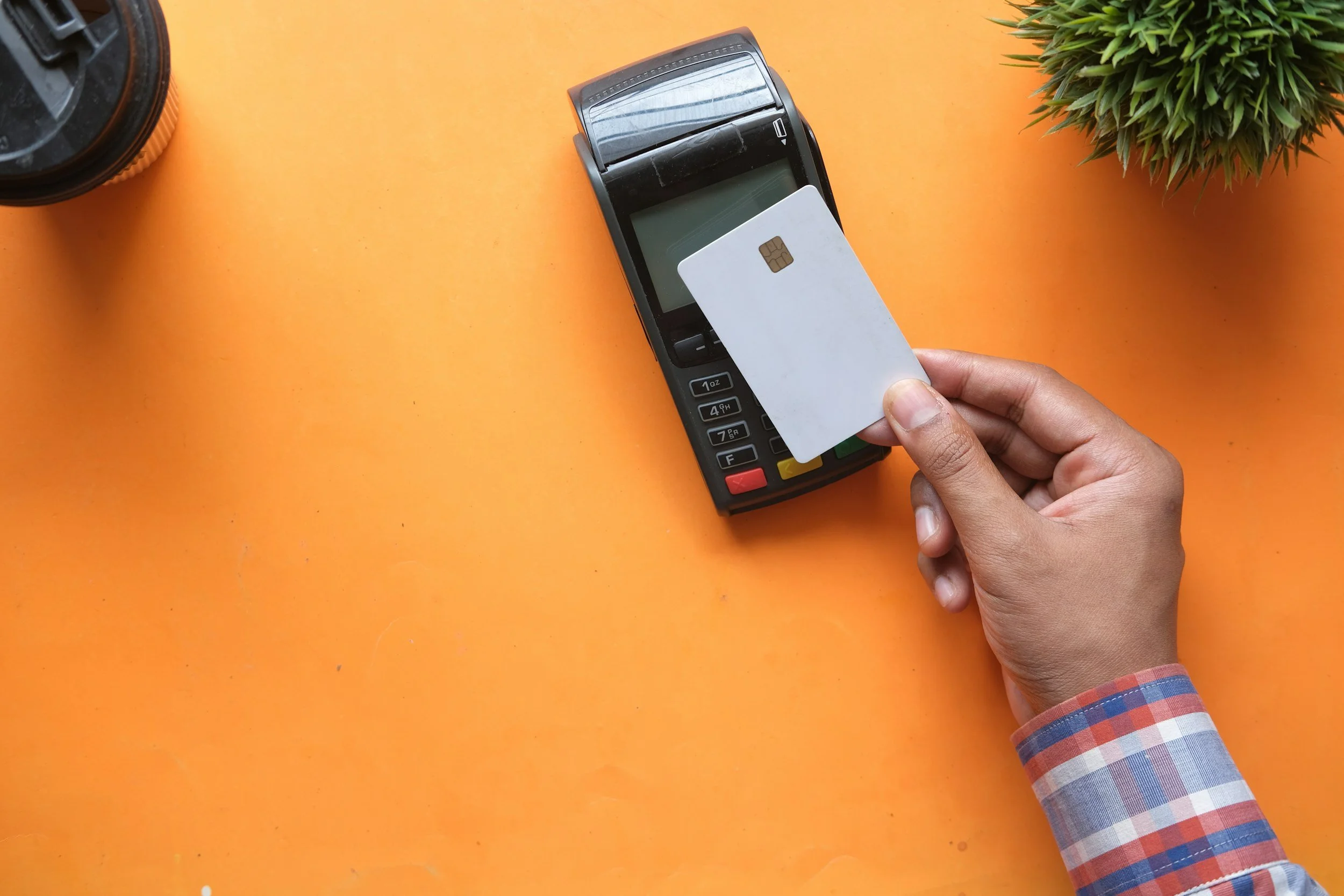 Person holding a credit card near a point-of-sale terminal on an orange surface, with a potted plant and a camera lens nearby.