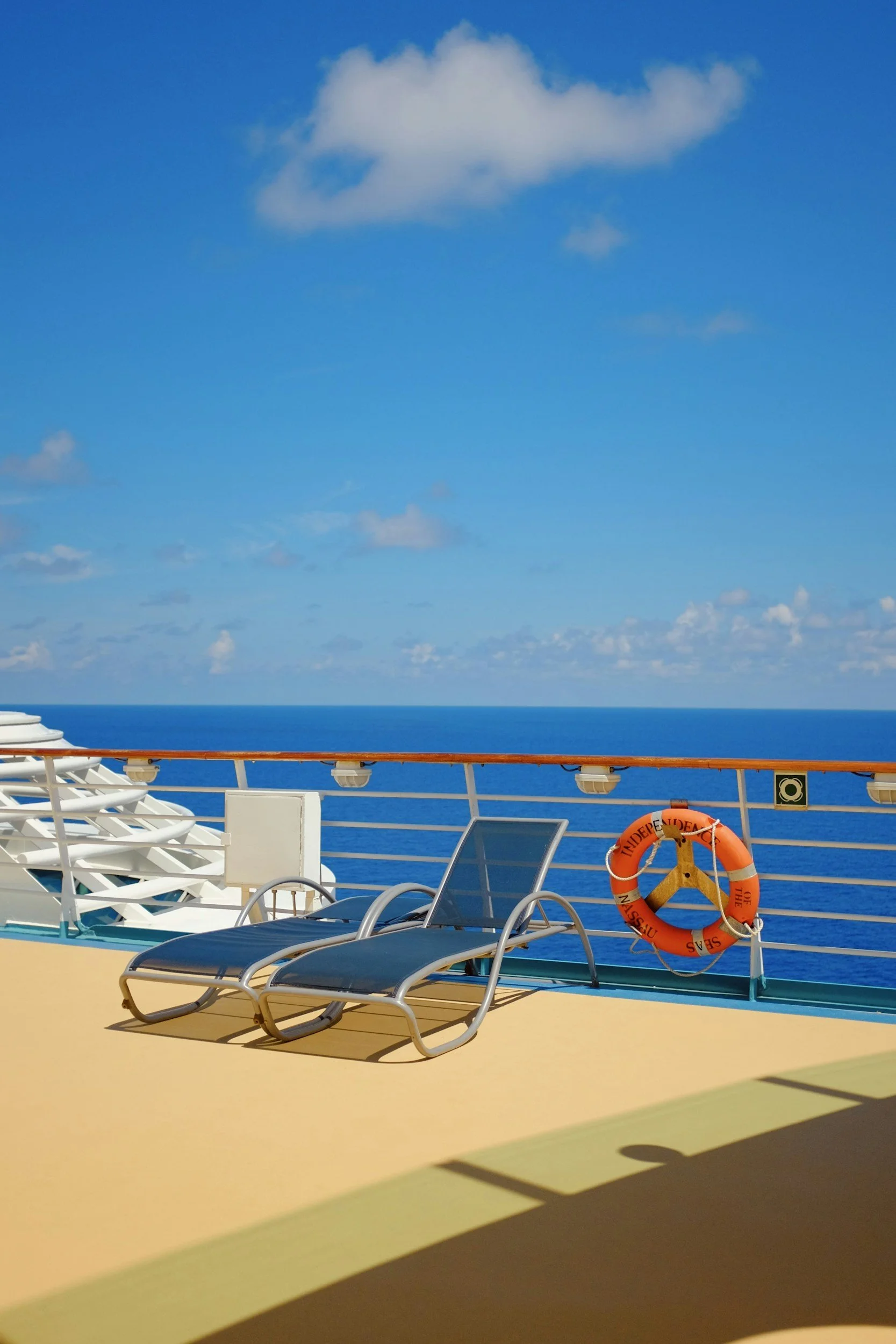 Deck chairs and a lifebuoy on the deck of a cruise ship with the ocean and a blue sky with clouds in the background.
