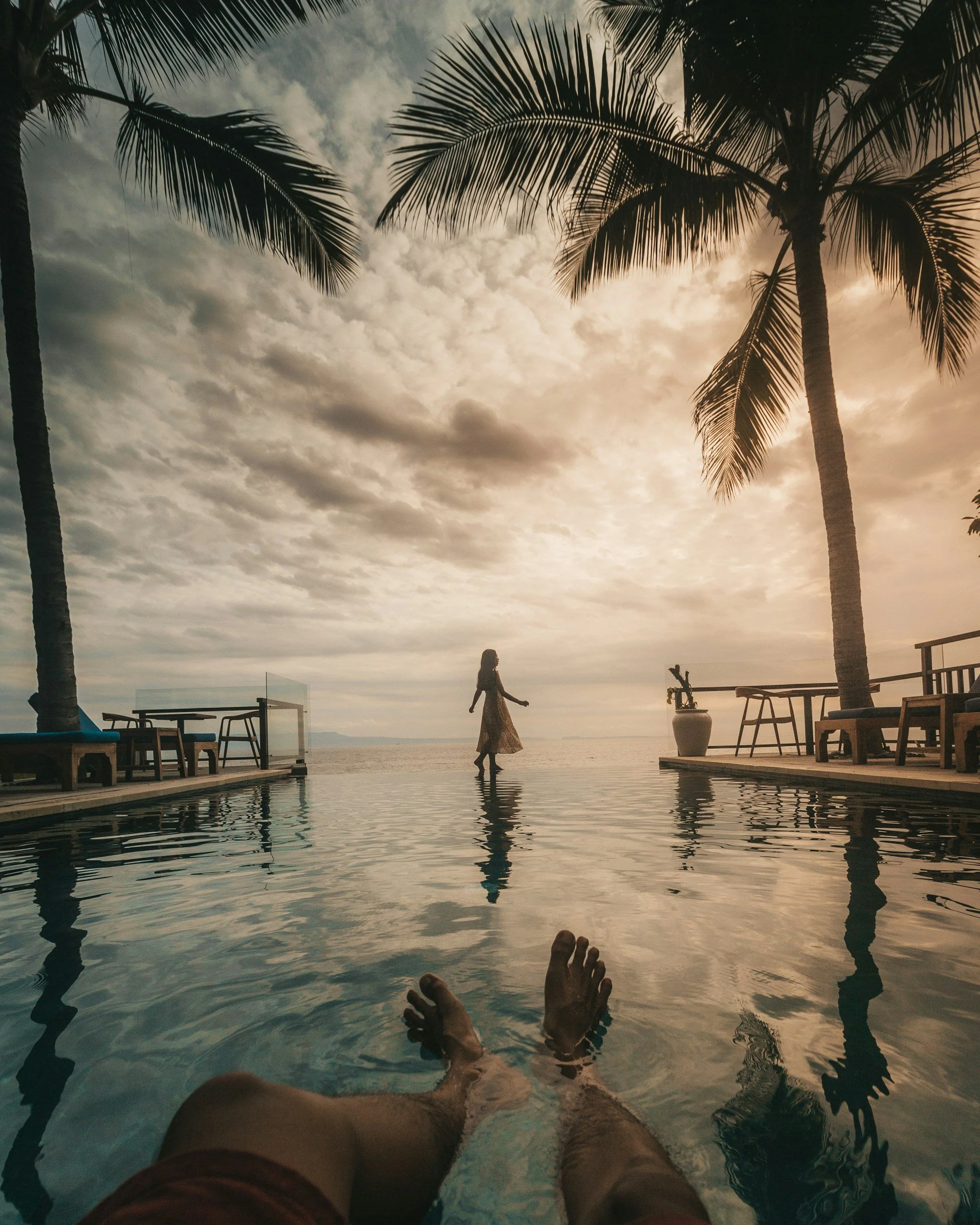A person is relaxing in a swimming pool with their legs extended towards the camera. The pool area is outdoors with palm trees, tables, and chairs. There is a person walking on the deck, and the sky is cloudy with a sunset or sunrise glow.