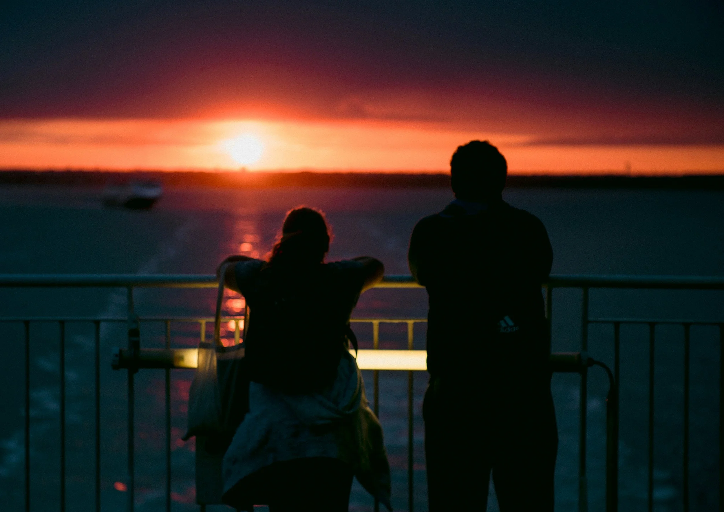 A couple, one sitting and one standing, watching a sunset over the ocean from a balcony.