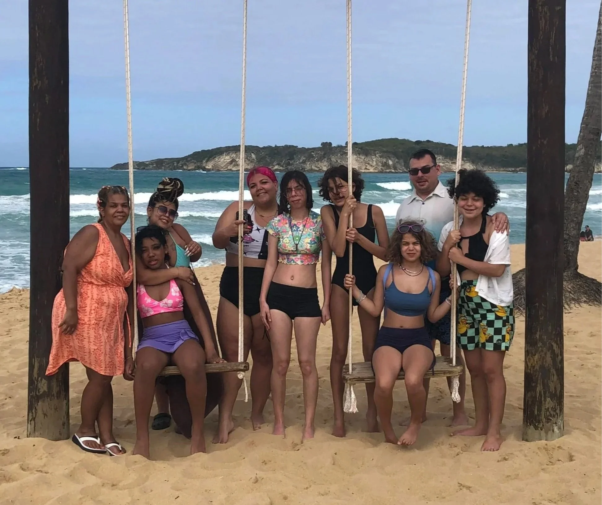 Group of people on the beach holding onto a wooden swing set framed by two large wooden posts, with a rocky island in the background and ocean waves.