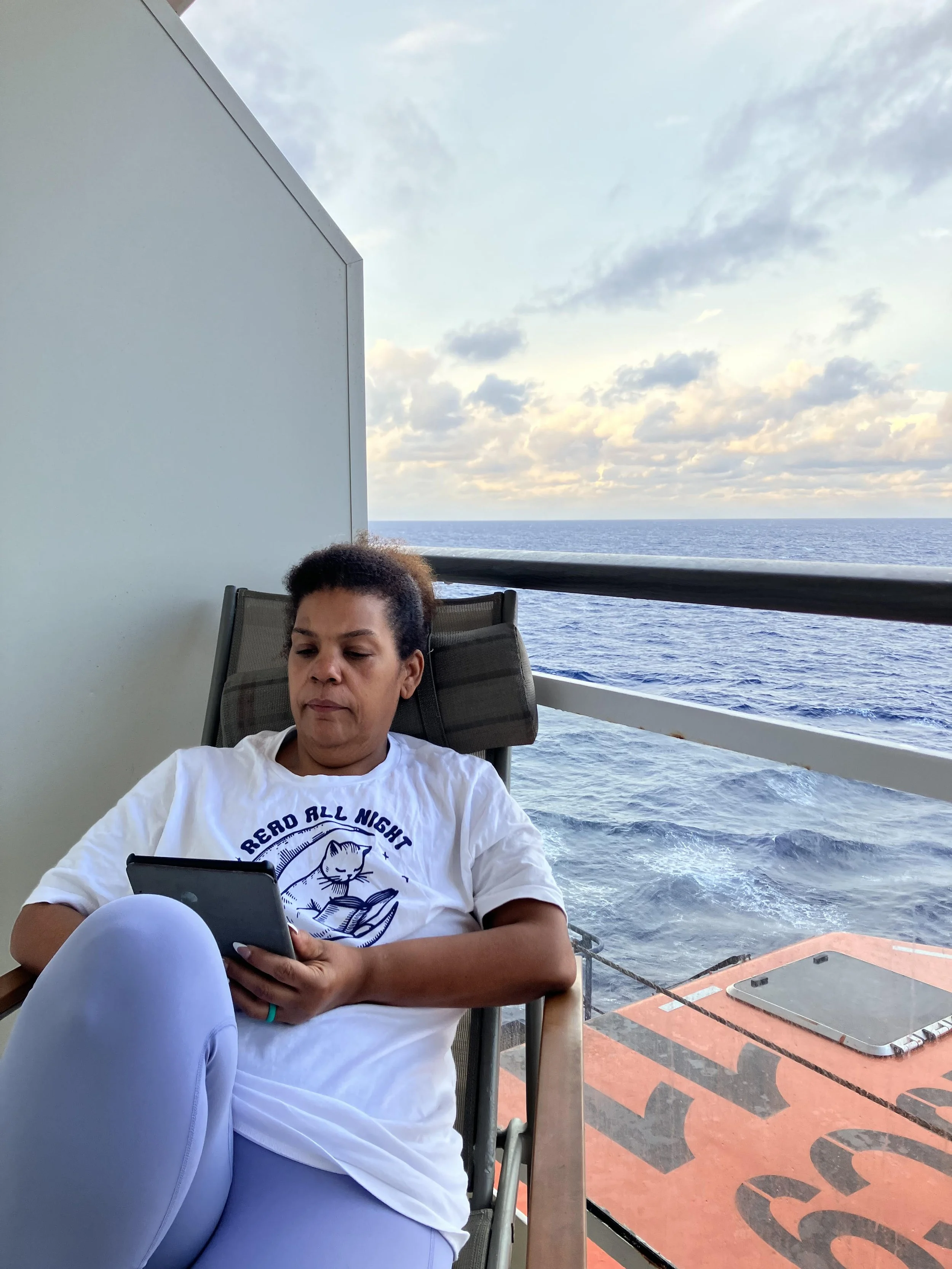 Person sitting on a boat deck, looking at their phone, with the ocean and a partly cloudy sky in the background.