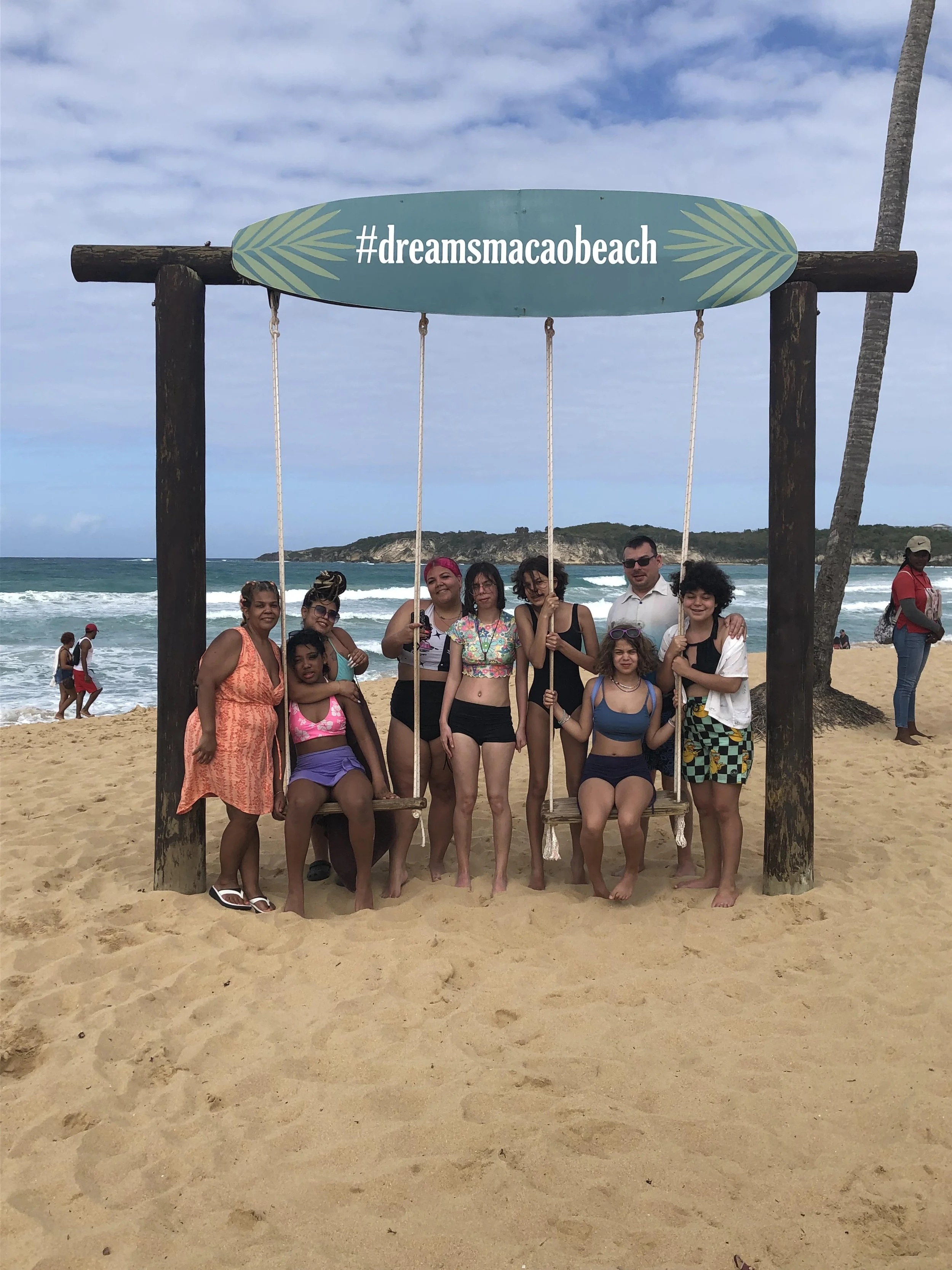 Group of people posing on a beach under a wooden swing with a sign reading '#dreamsmacaobeach'. The background shows ocean waves, a sandy beach, and a hilly landscape.