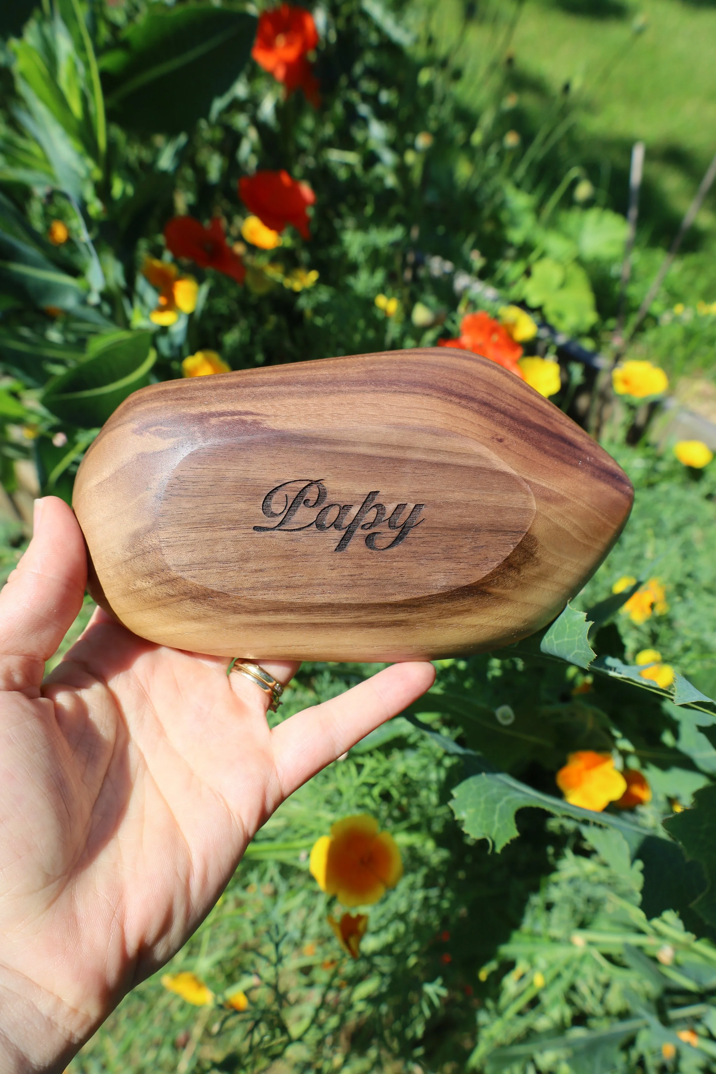 Une main tenant un bol en bois gravé avec le mot 'Papy', avec un jardin fleuri en arrière-plan.